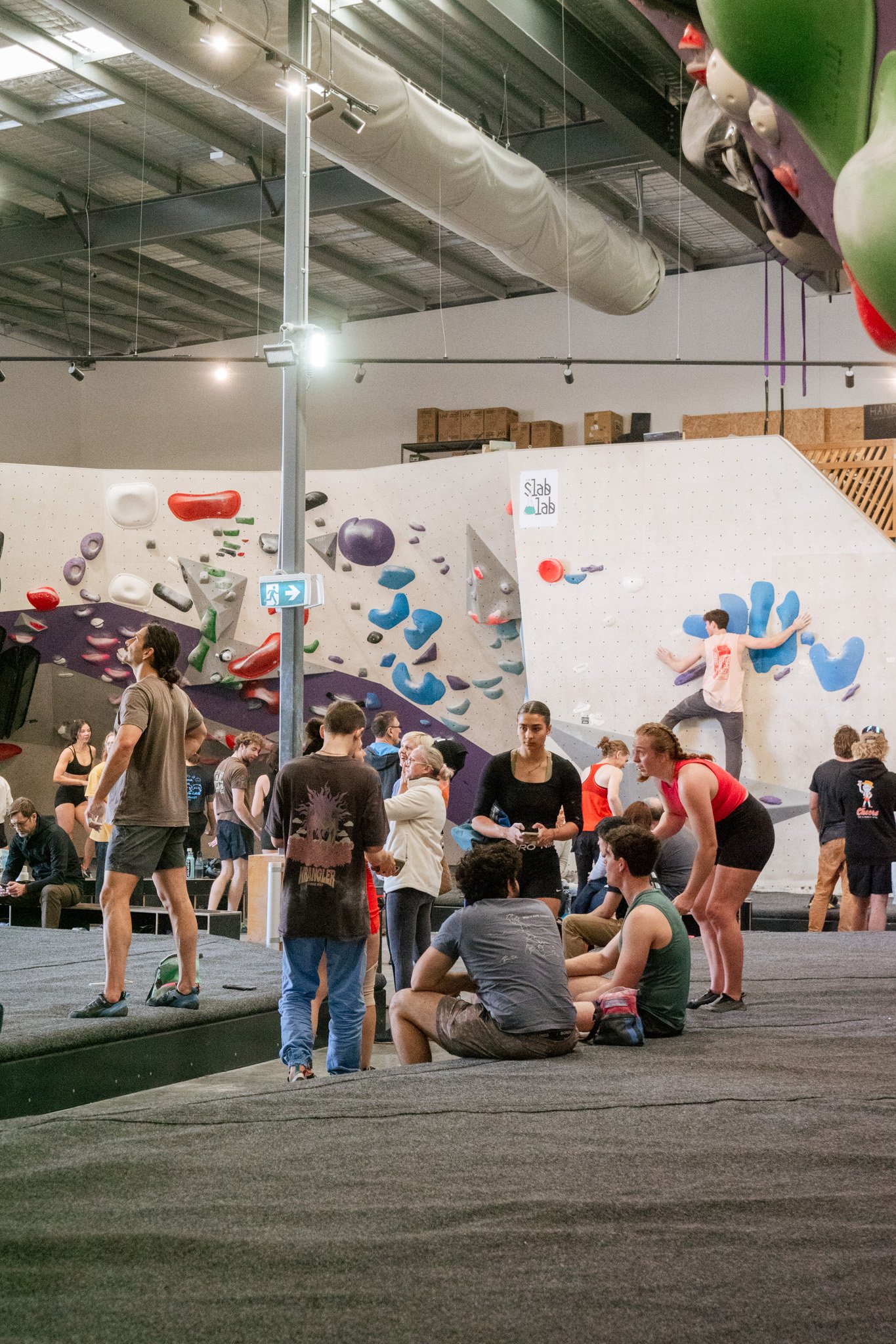 People at an indoor rock climbing gym, with several individuals bouldering on a wall covered with colorful climbing holds. Others are watching or talking nearby. The gym has high ceilings and industrial lighting.