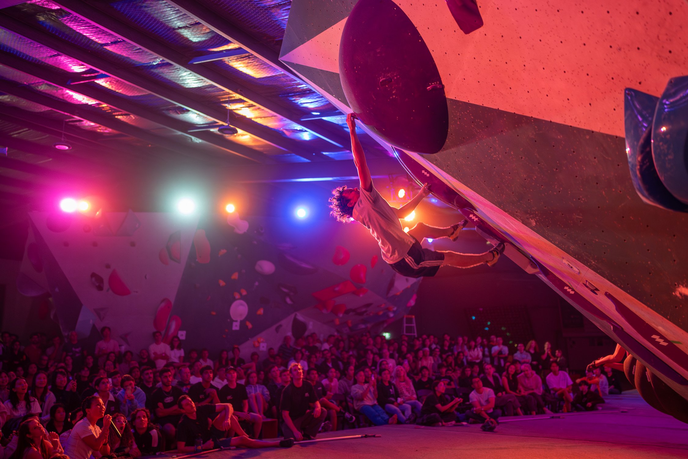 Indoor bouldering competition with climber on a rock wall, surrounded by an audience under colorful lighting.