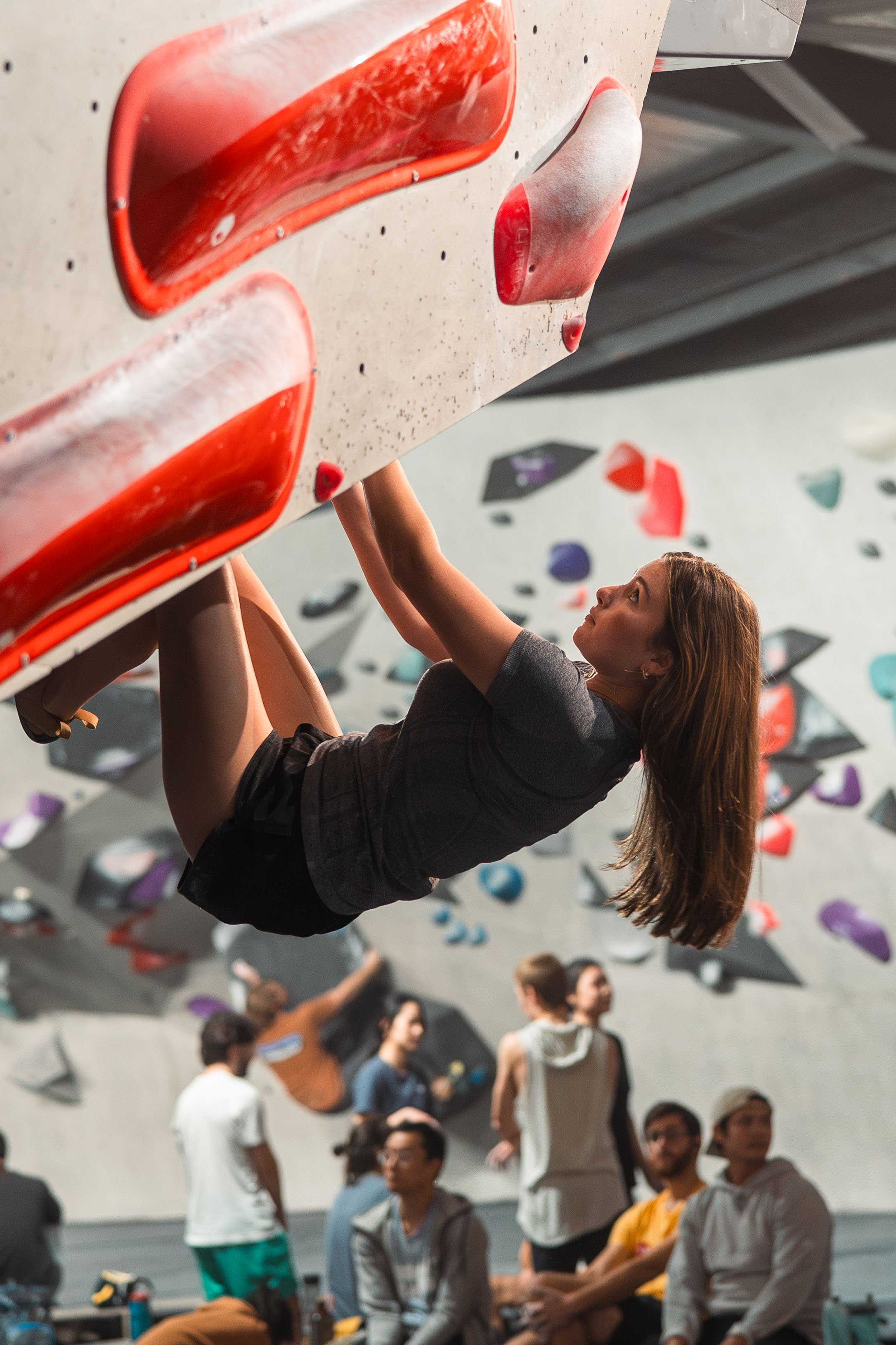 Woman bouldering on an indoor climbing wall with red holds, surrounded by other climbers in a gym setting.