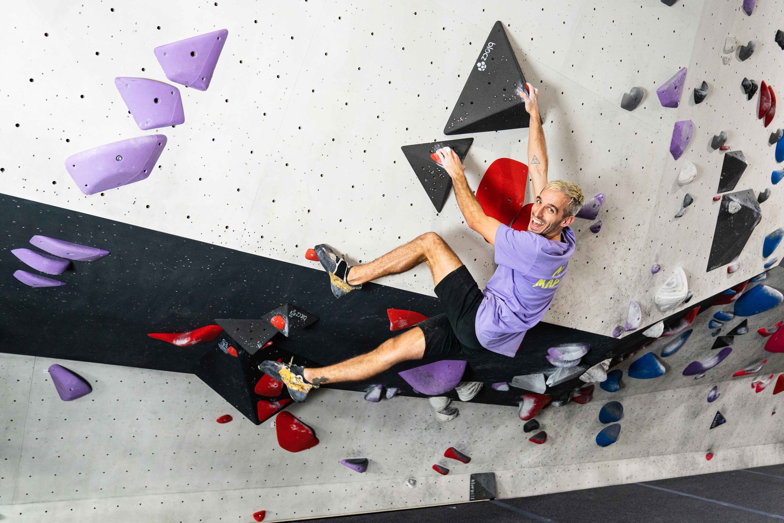 A man in a purple shirt rock climbing indoors on a climbing wall with colorful holds.