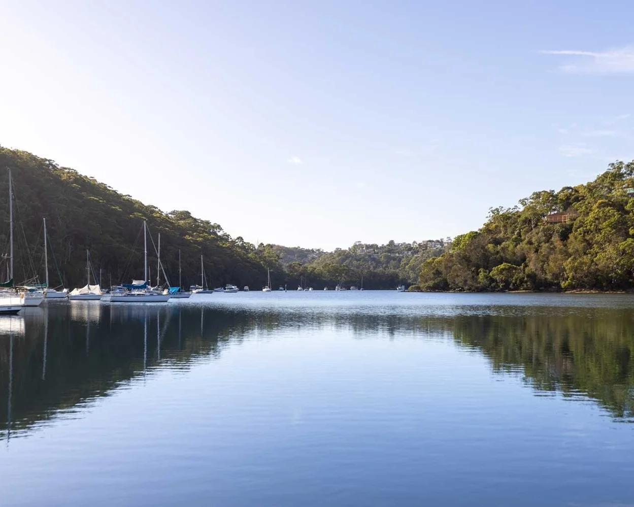 Scenic view of a tranquil bay with sailboats moored, surrounded by lush green hills, under a clear blue sky.