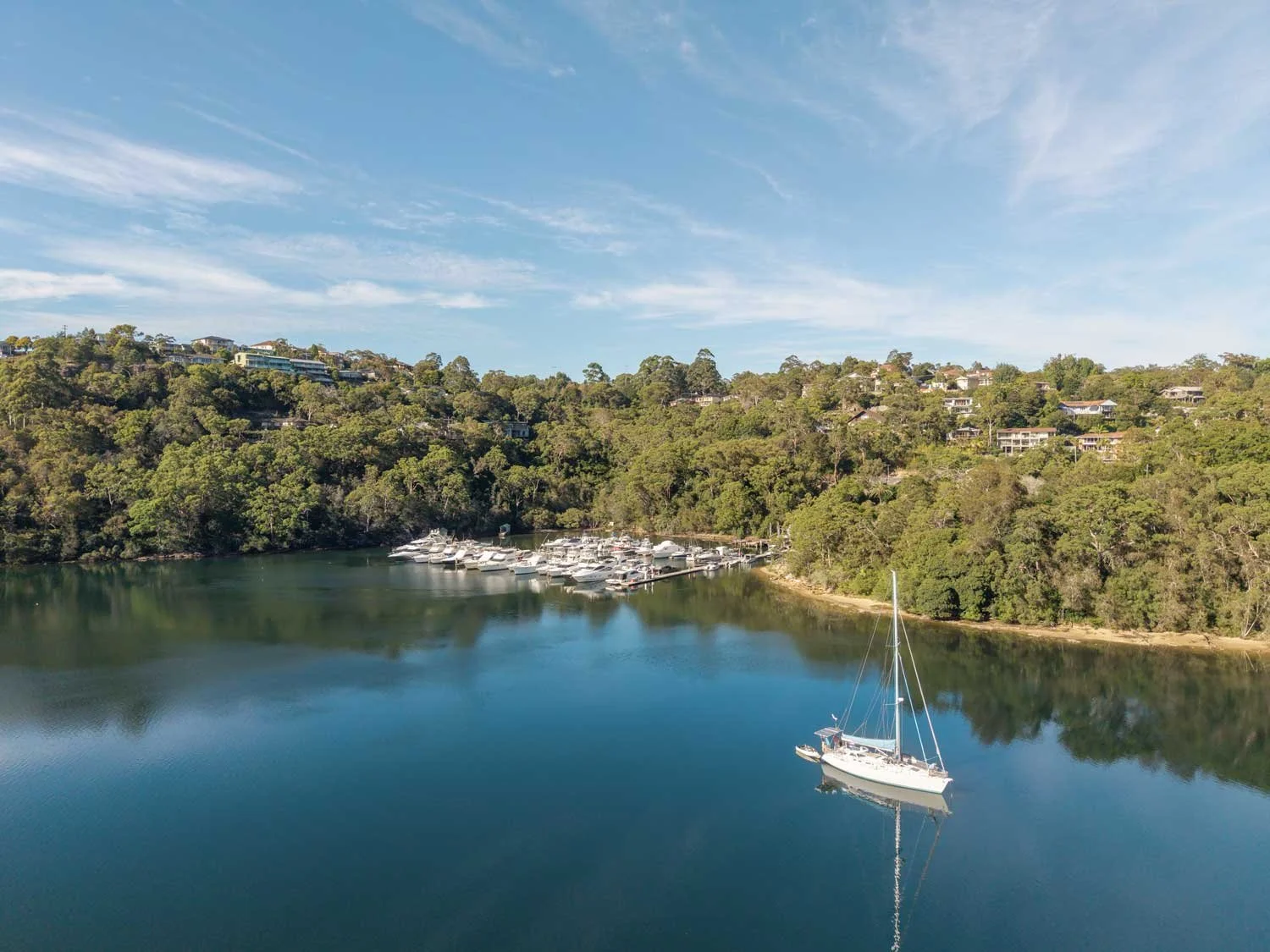Aerial view of a serene bay with a sailboat and a marina filled with yachts. Surrounding the bay are lush green trees and houses on a hillside under a clear blue sky.
