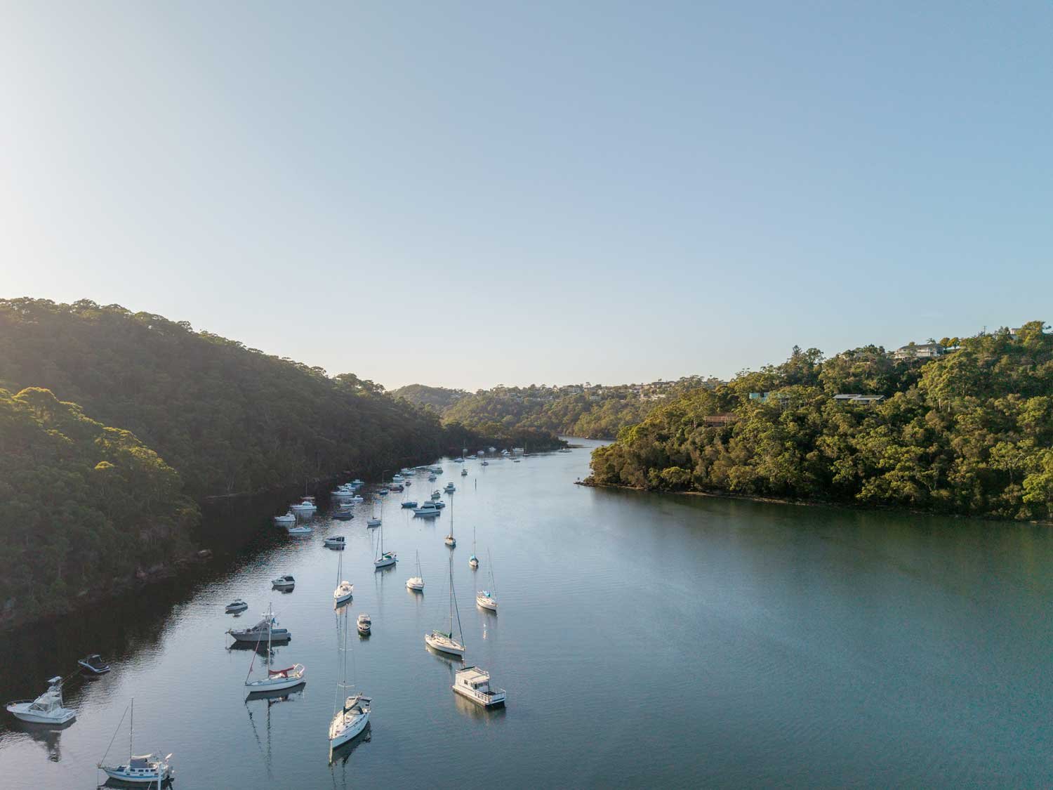 Aerial view of a serene river surrounded by lush green hills with numerous boats anchored in the water.