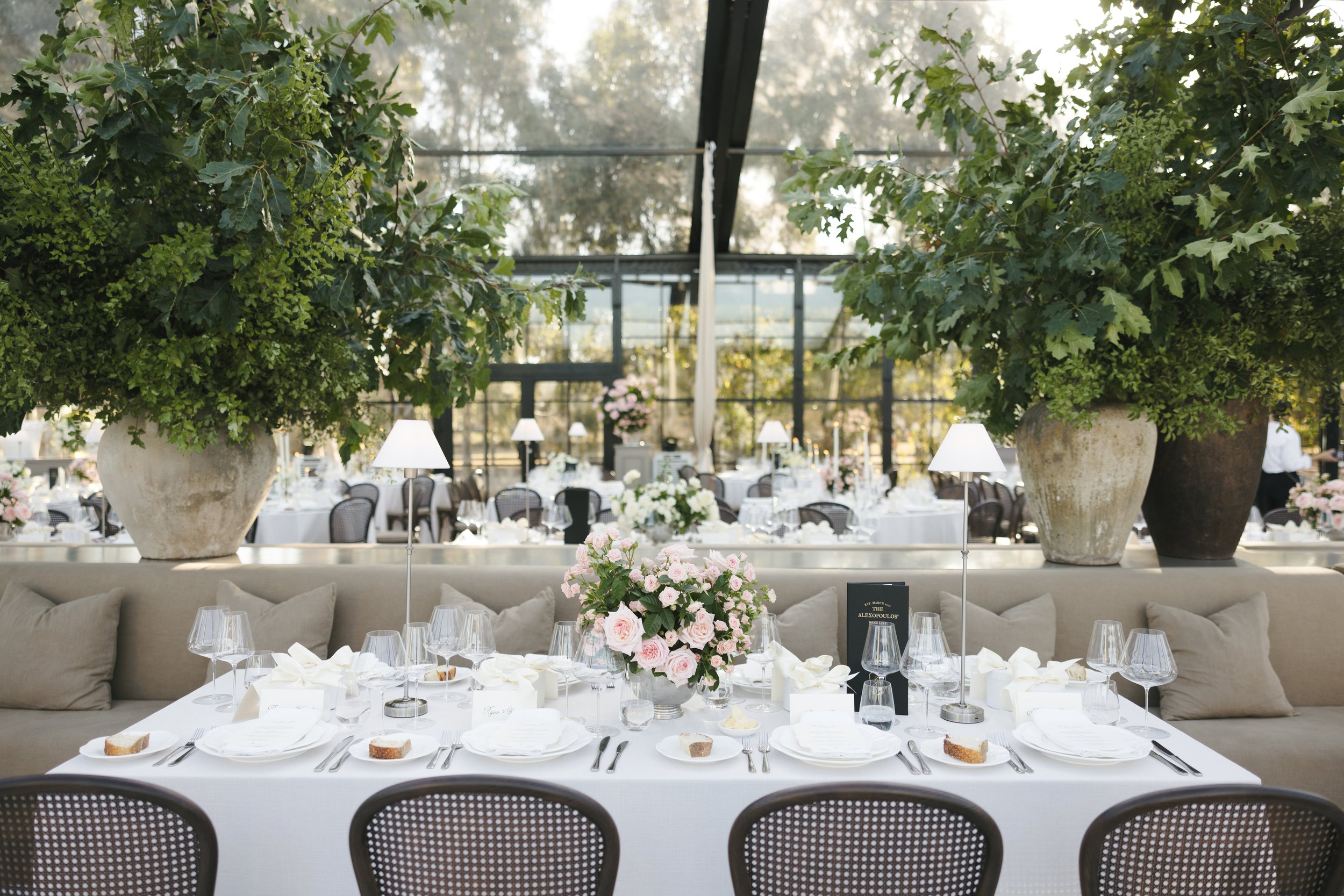 Elegant outdoor dining setup with white tablecloths, floral centerpieces, large potted plants, wine glasses, and tableware, under a glass roof in a garden setting.
