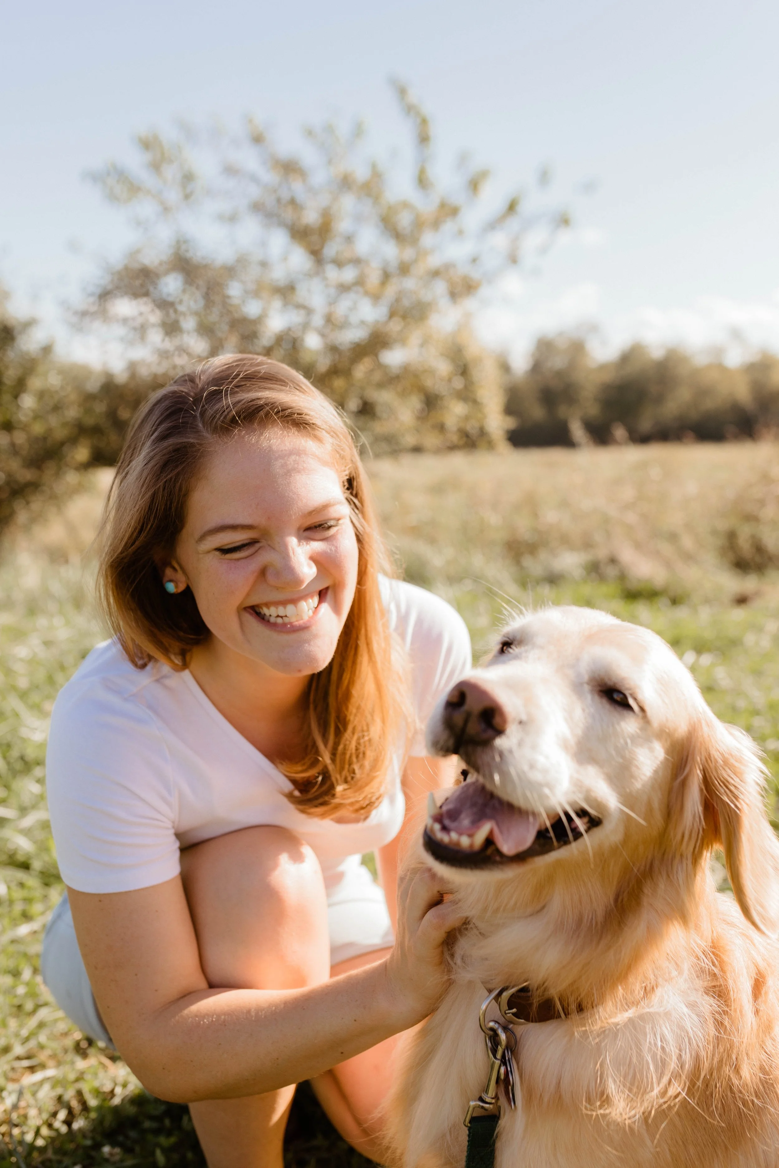 A woman with red hair smiling and petting a happy golden retrieverdog outdoors on a sunny day with trees and grass in the background.