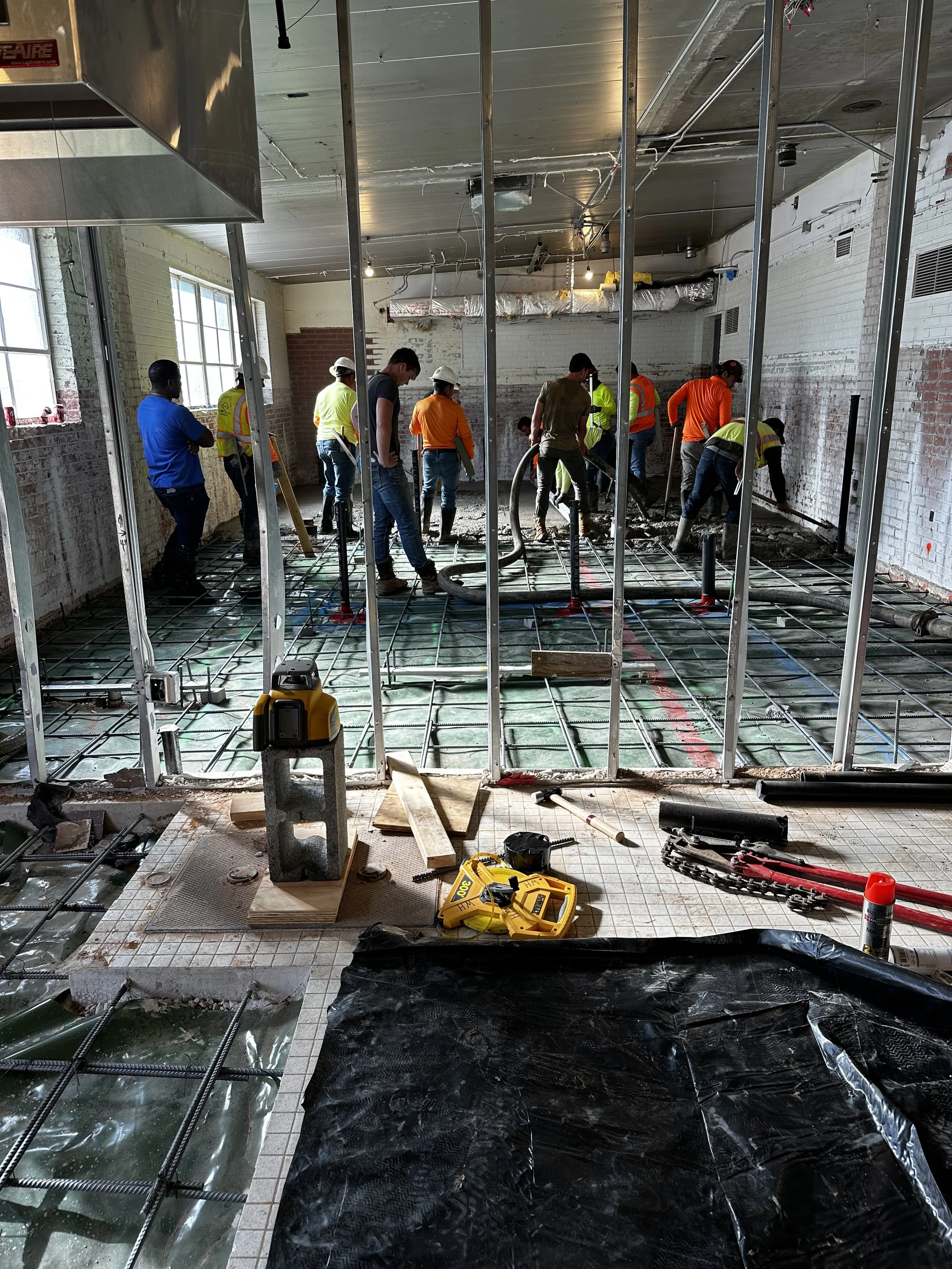 Construction workers pouring concrete inside a building with exposed brick walls and large windows, tools and equipment scattered on the floor.