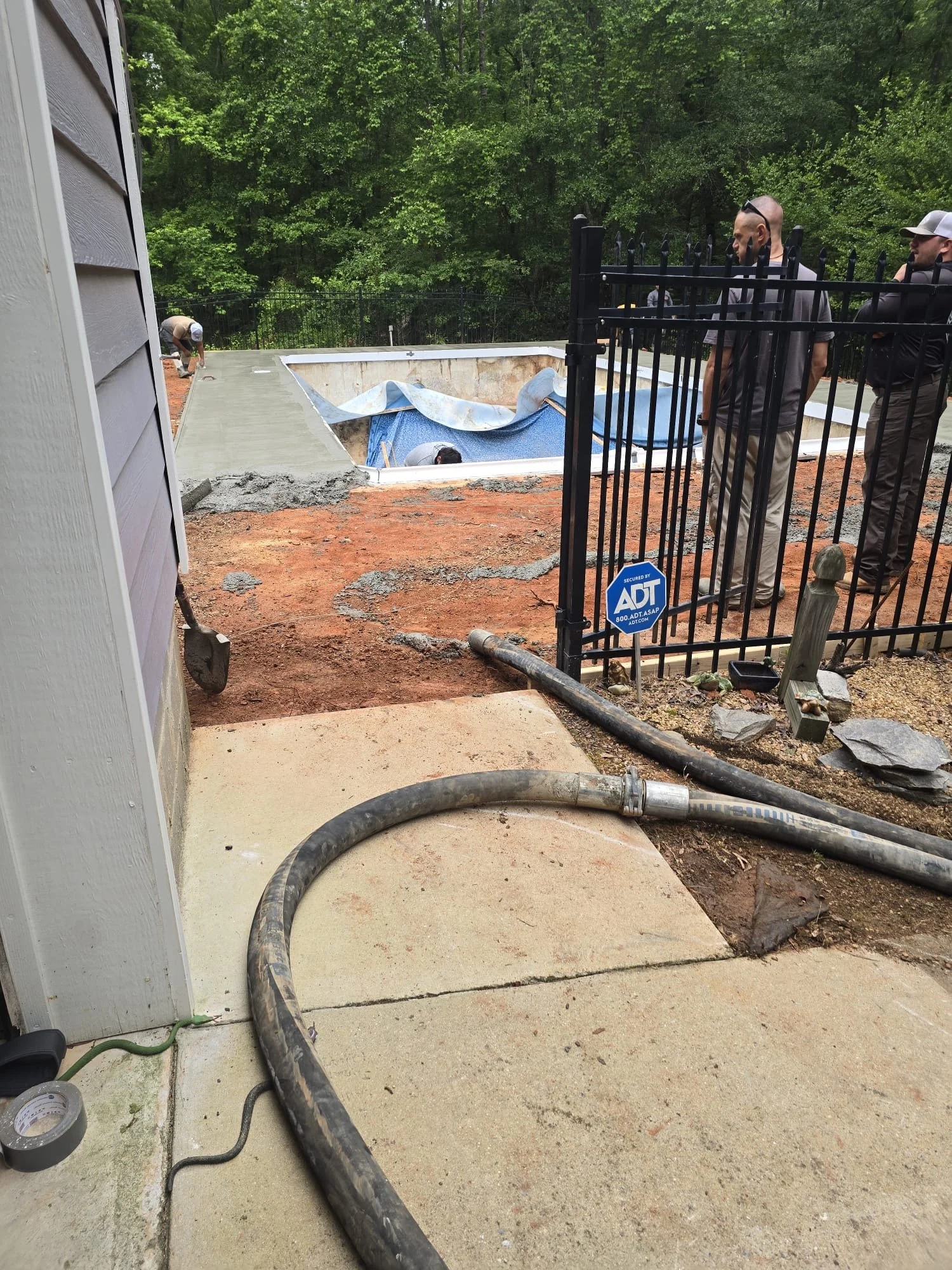 Workers installing a new pool in the backyard with a concrete deck, surrounded by trees and a black metal fence.