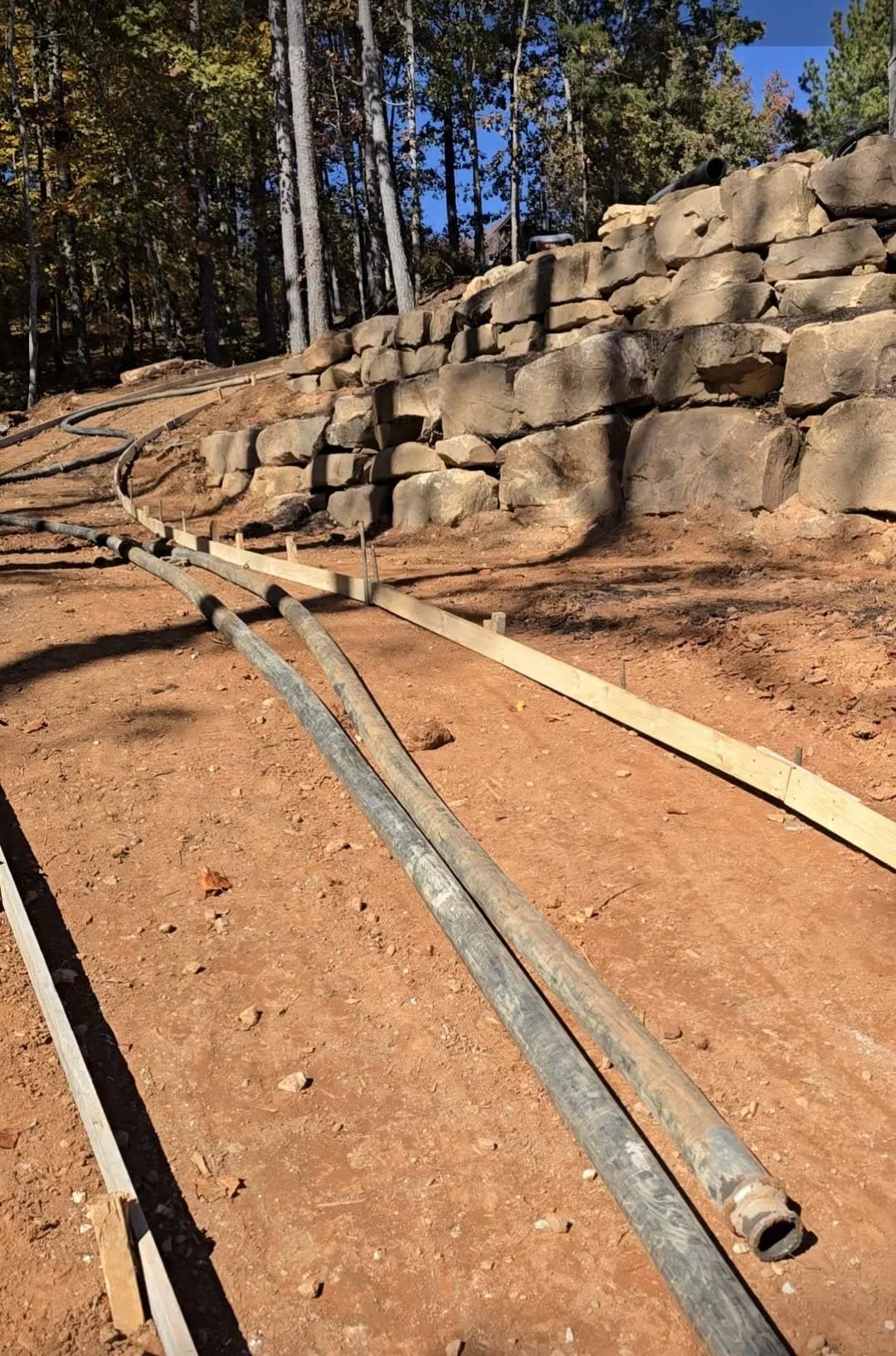 Construction site with bent pipes laid on the ground, a wooden barrier, and a large stone retaining wall in the background amid trees.