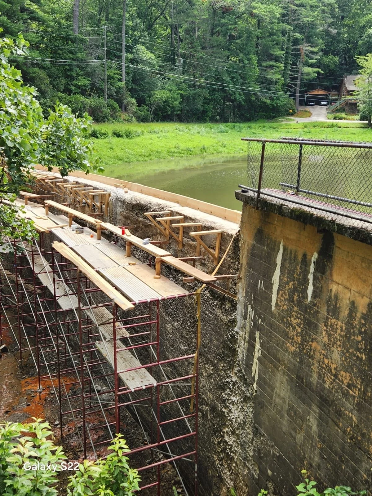 Construction of a retaining wall with wooden support beams and metal scaffolding along a riverbank, surrounded by green trees and grass.