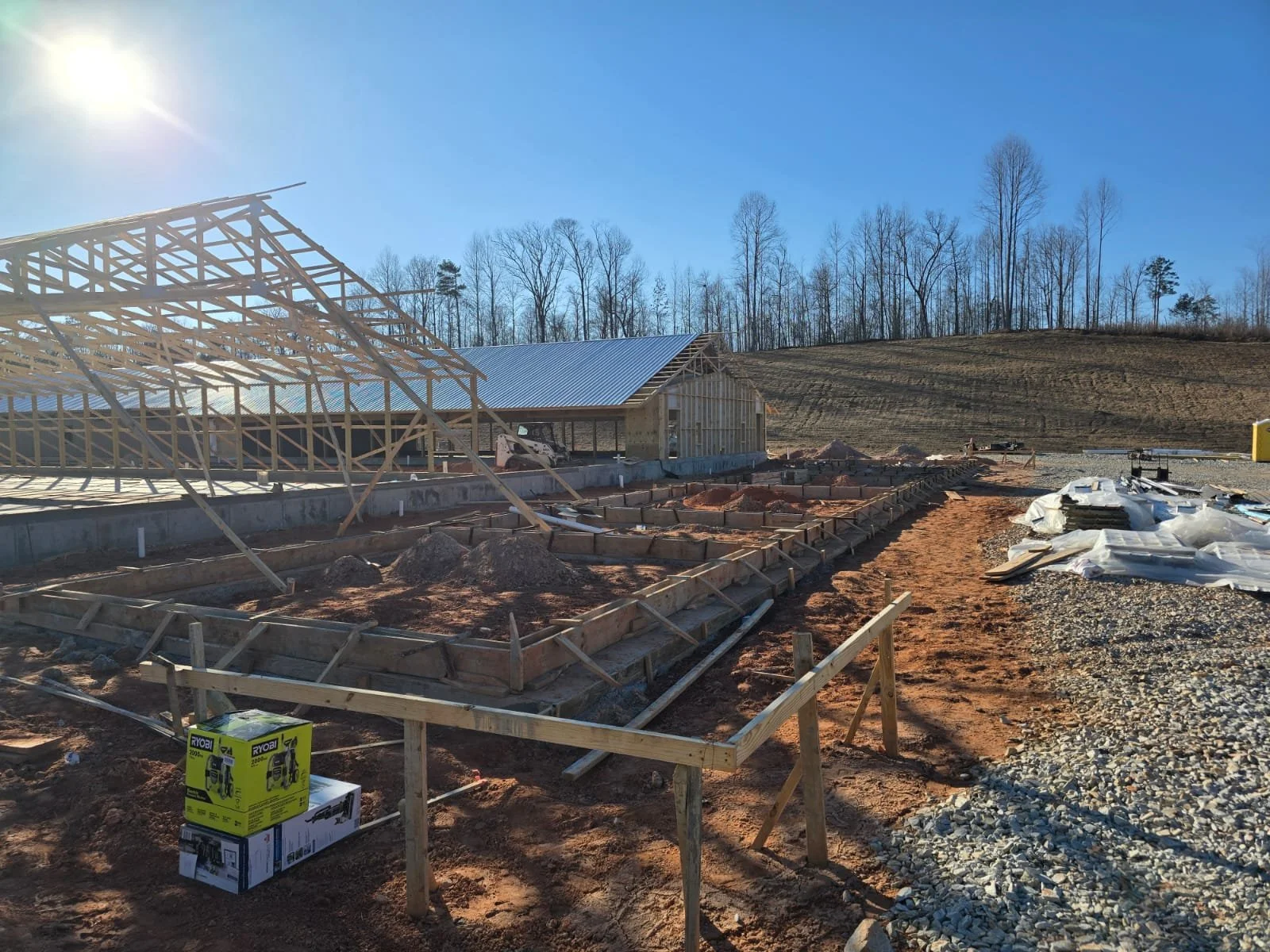 Construction site with wooden framing and framework for a building, dirt and gravel ground, construction materials and tools, and trees in the background under a clear blue sky with sunlight.