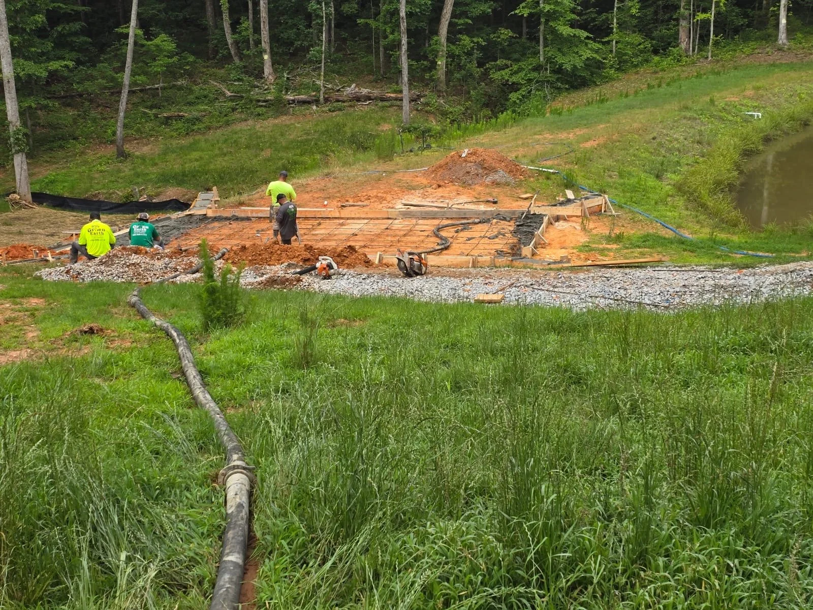 Construction site with workers pouring concrete foundation, surrounded by green grassy area and wooded hills.