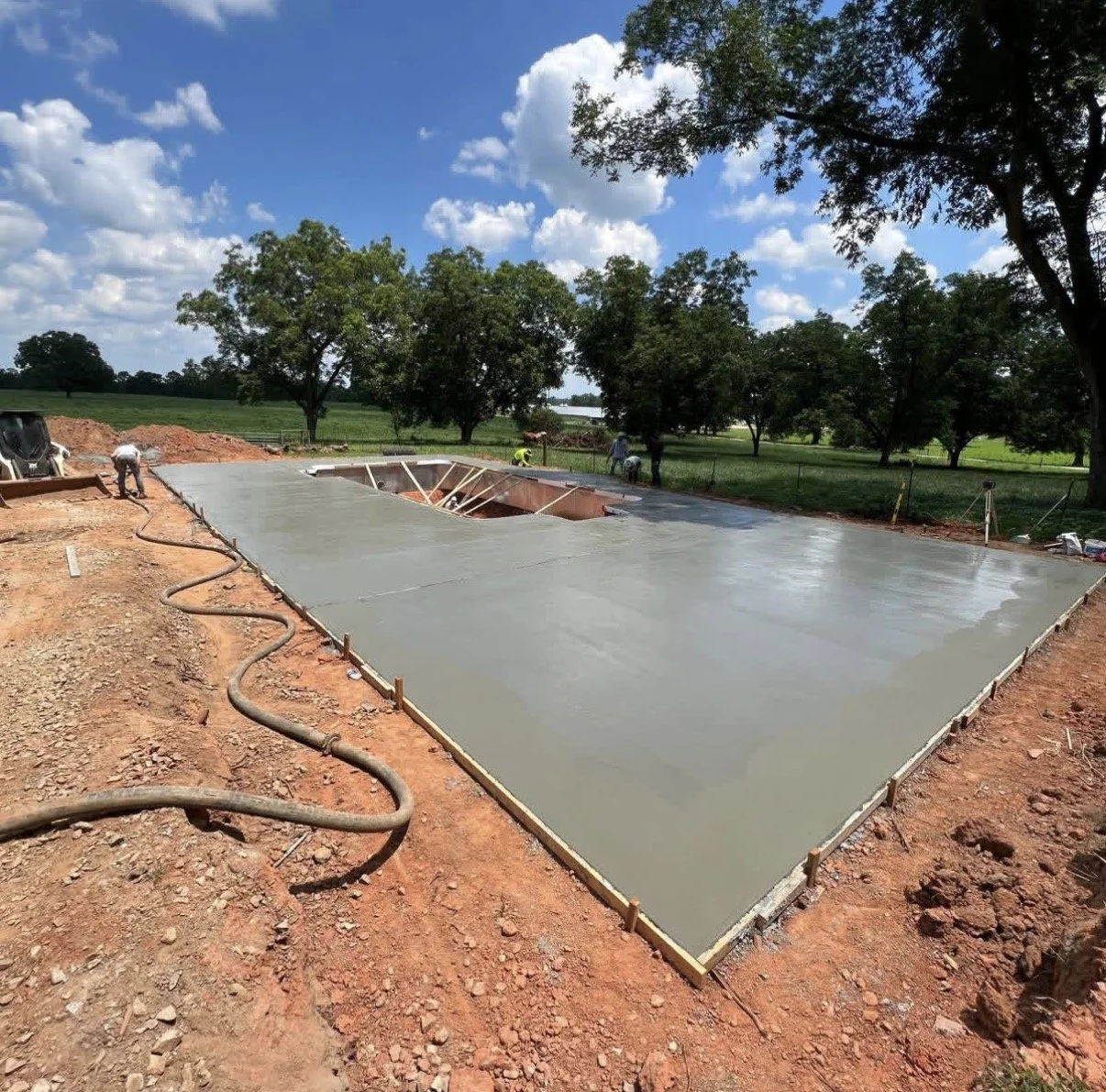 Construction workers pouring and leveling a concrete slab foundation outdoors on a sunny day with trees and green fields in the background.