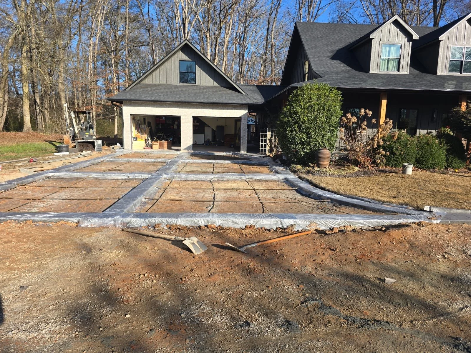 A house under renovation with a concrete driveway being poured and finished, surrounded by trees, with work tools visible in the garage.