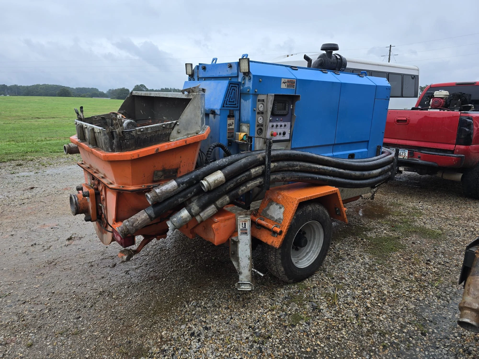 A street-cleaning machine with a blue body, orange parts, and black water hoses is parked on a gravel surface. Behind it, there are two vehicles, including a red truck and a white bus, with a green field and cloudy sky in the background.