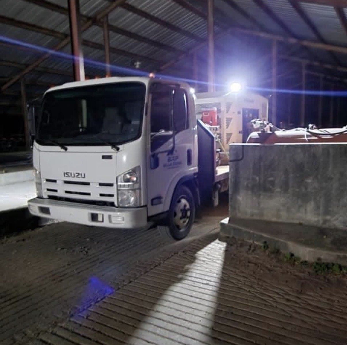 A white Isuzu flatbed truck parked inside a large warehouse or garage with a corrugated metal roof, illuminated by bright lights overhead.