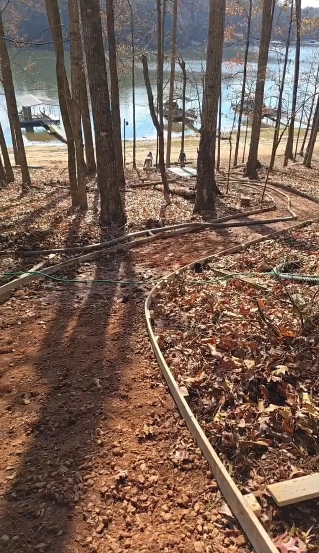 A dirt path in a wooded area leading to a lake, with some of the ground covered in fallen leaves. There are trees with tall, thin trunks and shadows cast on the ground. A wooden dock extends into the lake in the background.