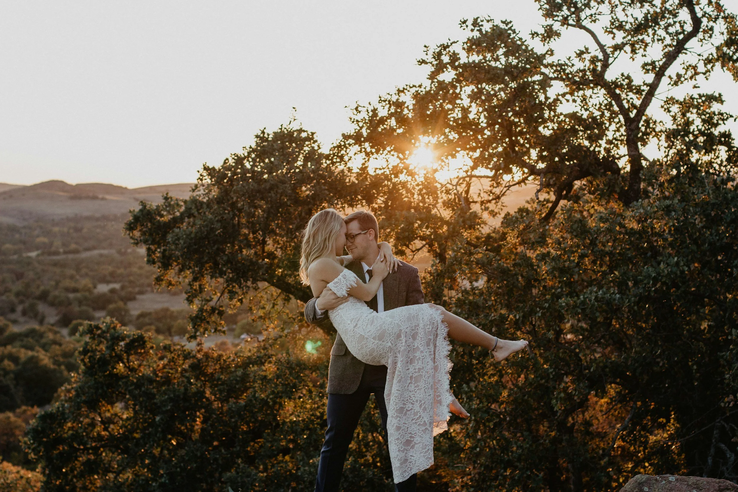 A man holding a woman in a white lace dress during sunset, with trees and hills in the background.