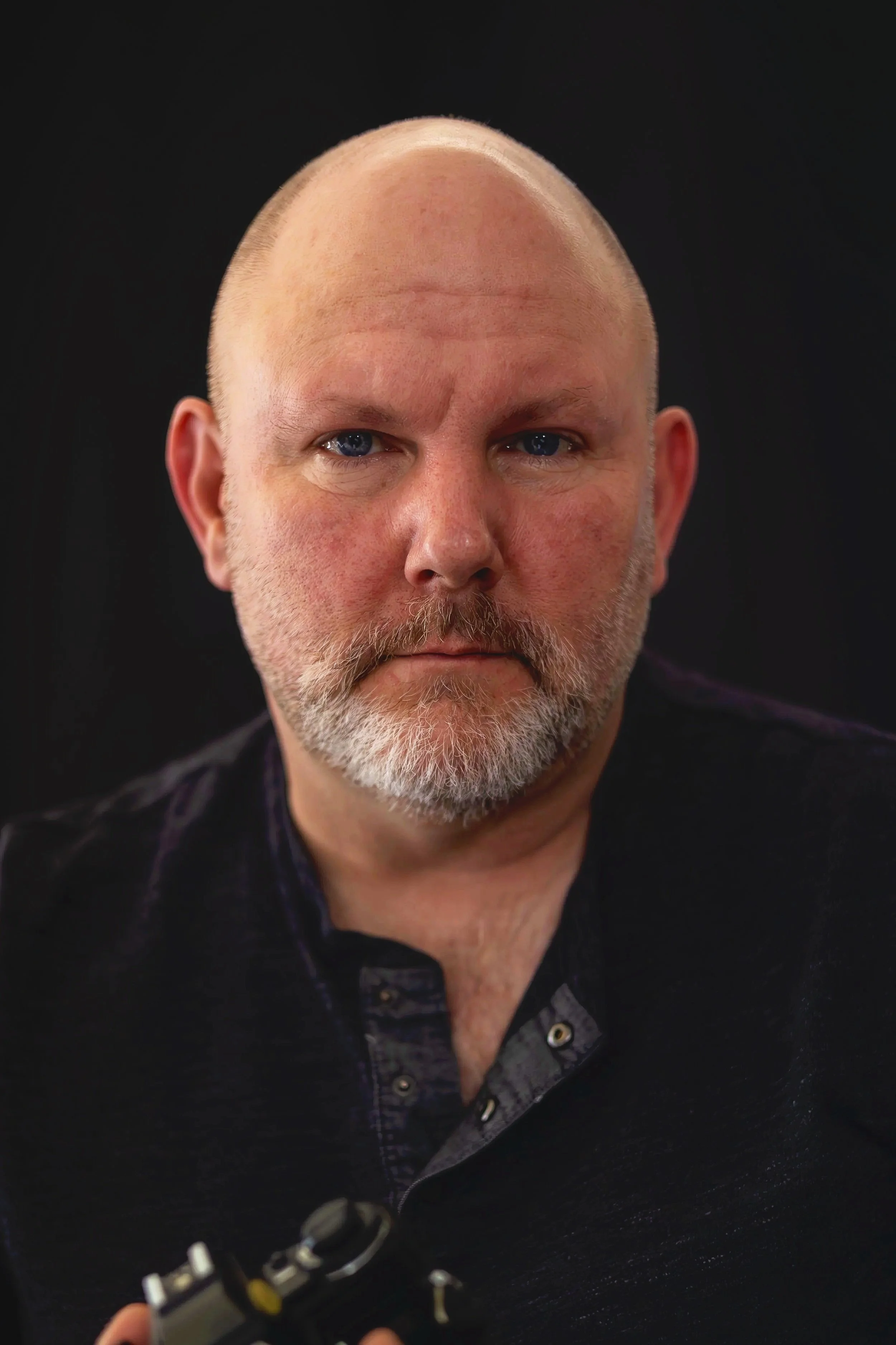 A close-up portrait of a middle-aged man with a bald head, blue eyes, and a beard, wearing a dark shirt with a slightly open collar, against a black background.