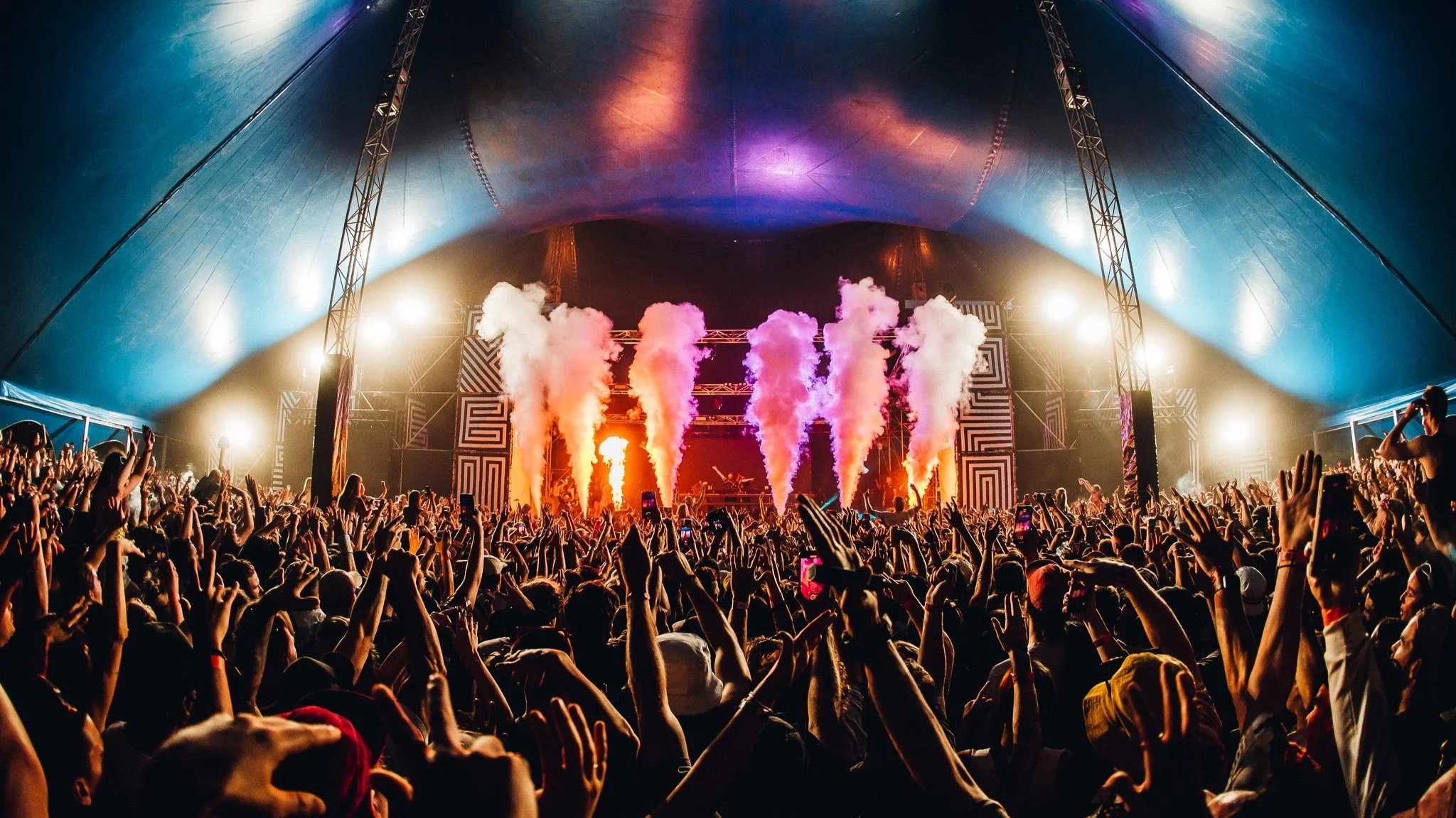 Crowd at a concert with stage lights and pyrotechnics inside a large tent.