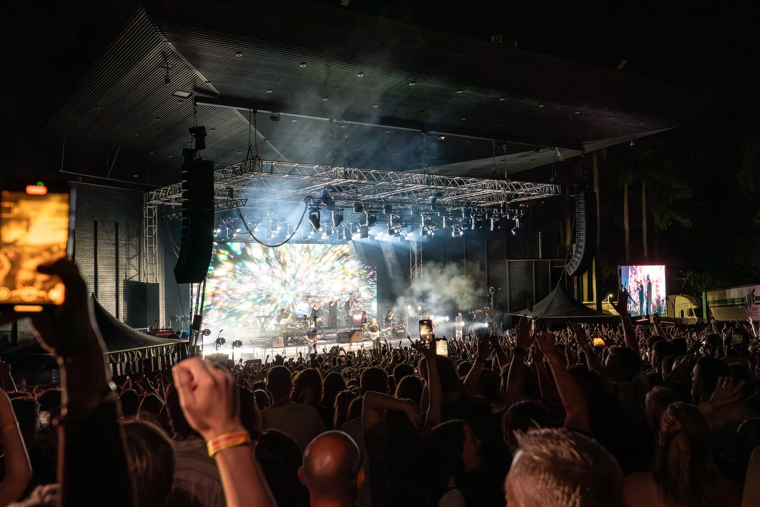 A large crowd watching a live outdoor concert at night, with a brightly lit stage, musicians, and a colorful digital background on screens.