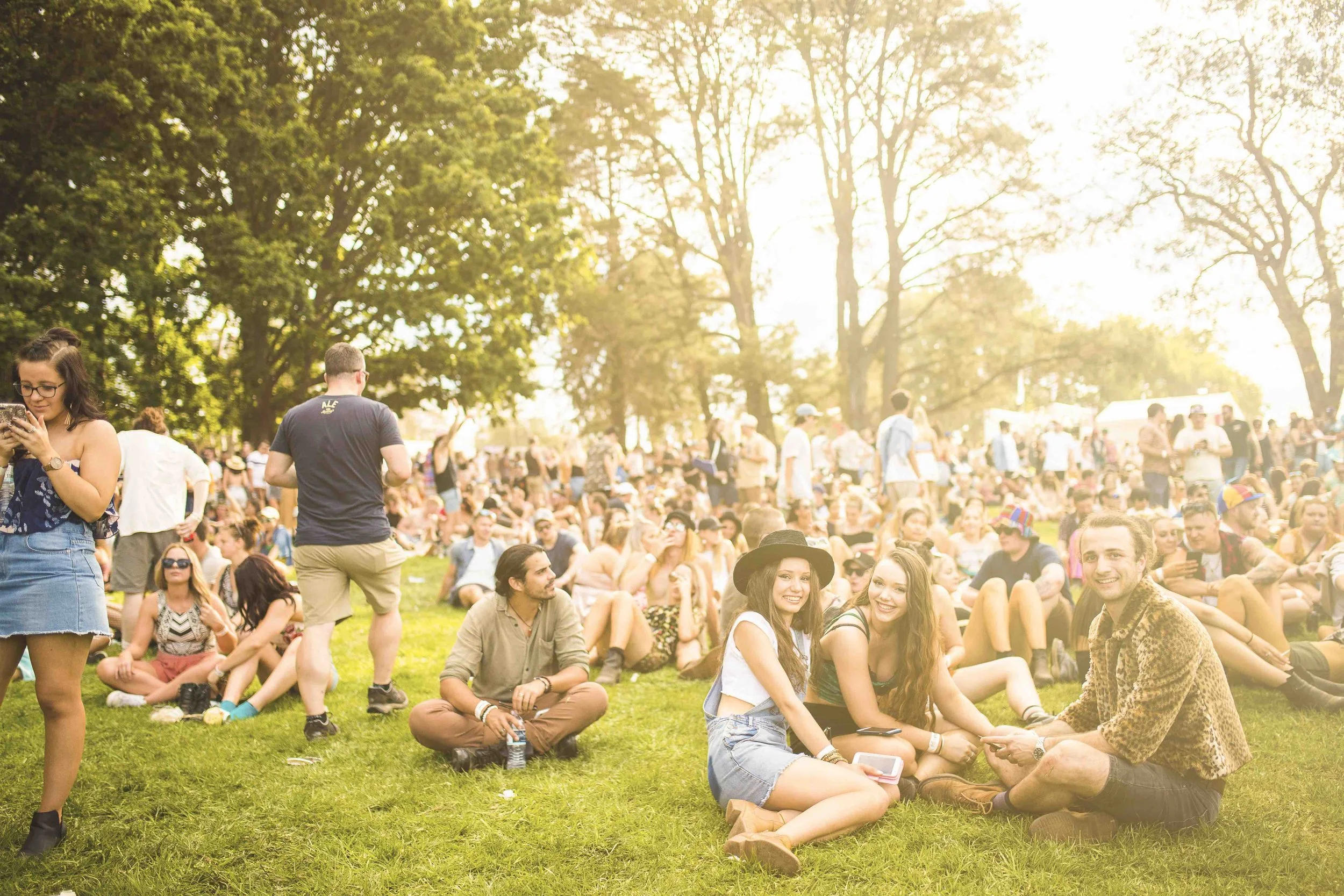 A large group of people gathered outdoors on a grassy area, many sitting or lying on the grass, with some standing and walking. The scene is sunny with bright sunlight filtering through trees in the background, indicating a cheerful outdoor event or festival.