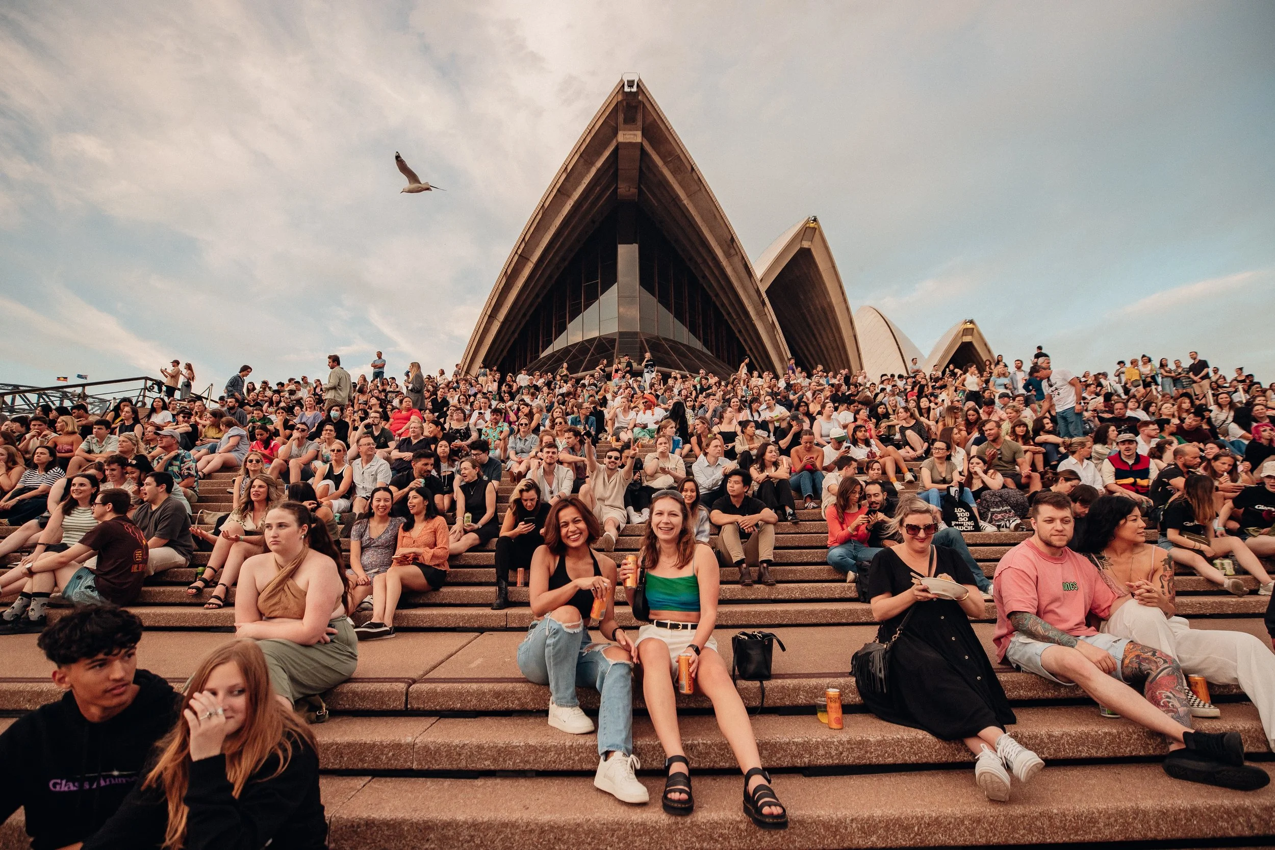 Crowd of people sitting on steps outside the Sydney Opera House during sunset, with some holding drinks and others using their phones