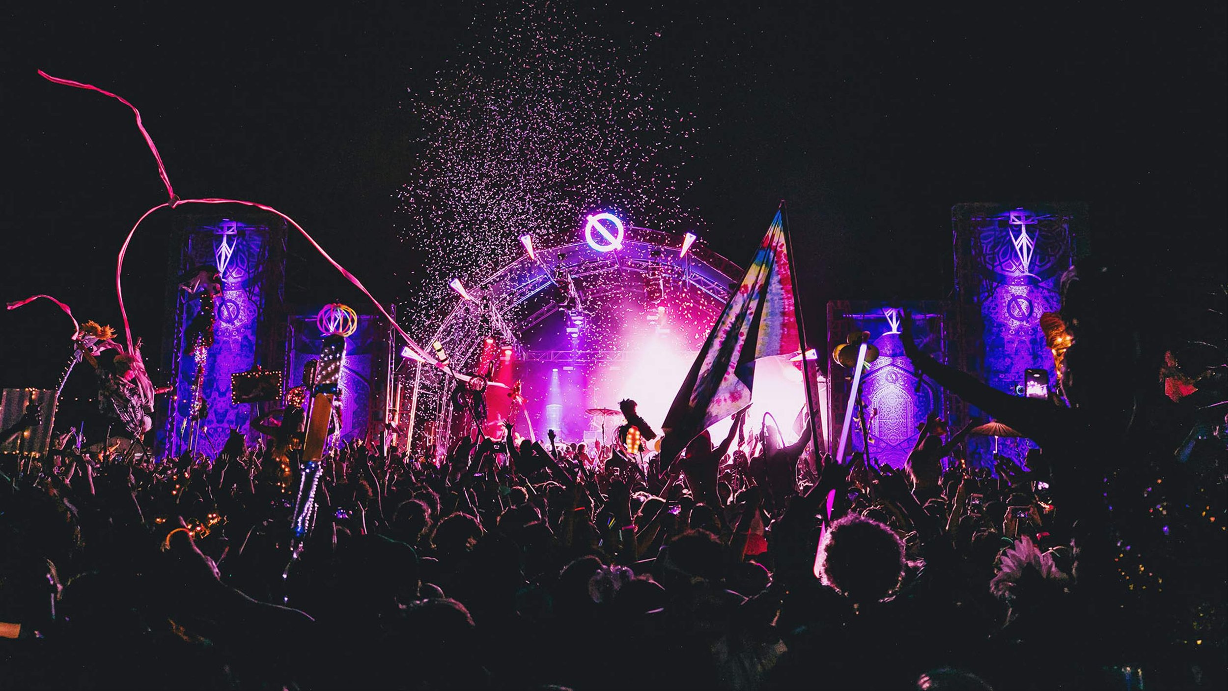 A night concert scene with a large crowd, colorful lights, and fireworks. The stage is lit with purple and pink lights, and flags are being waved by the audience.