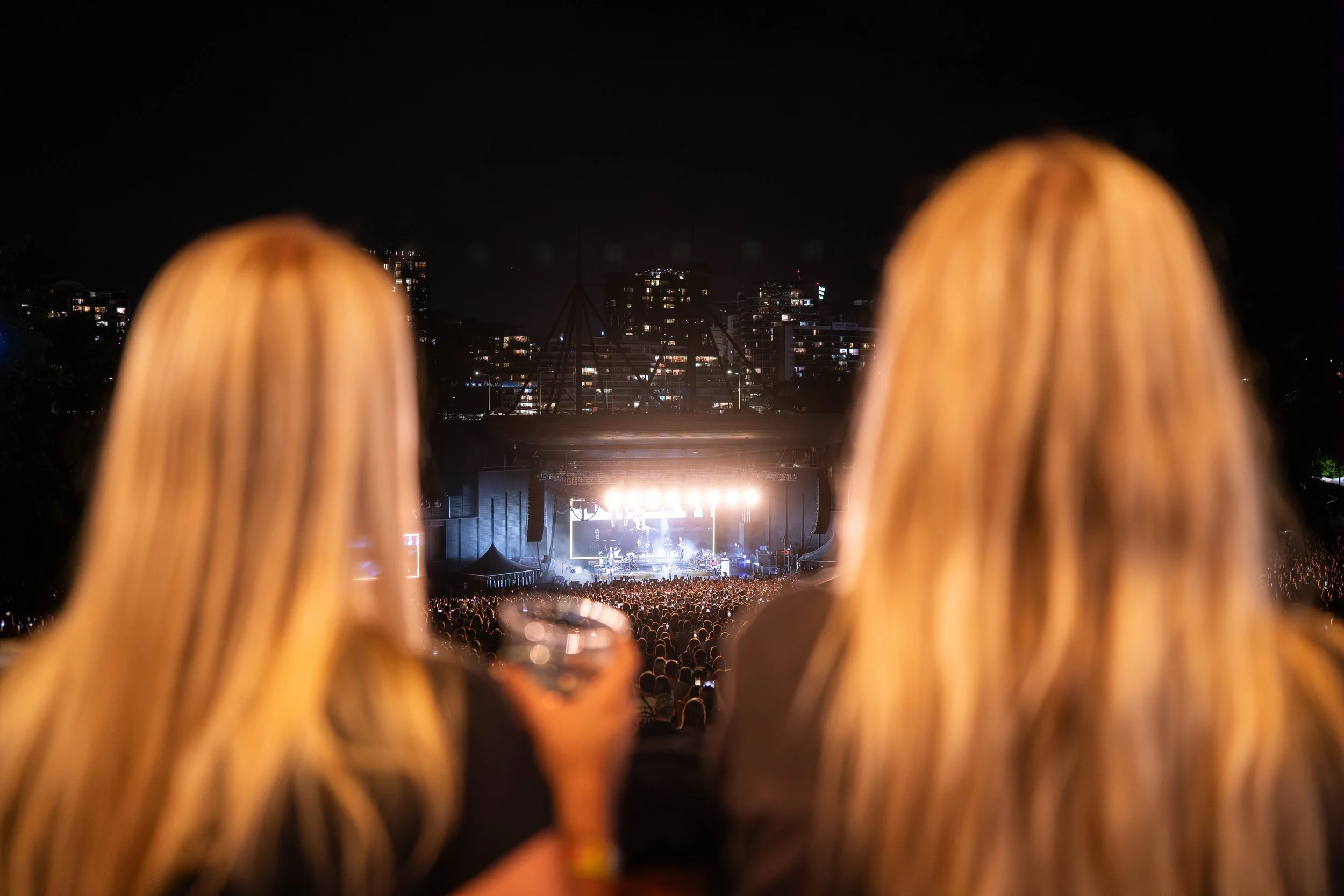 Two women with long blonde hair watching a concert at night, with the stage lit up and a large crowd in front, city buildings in the background.
