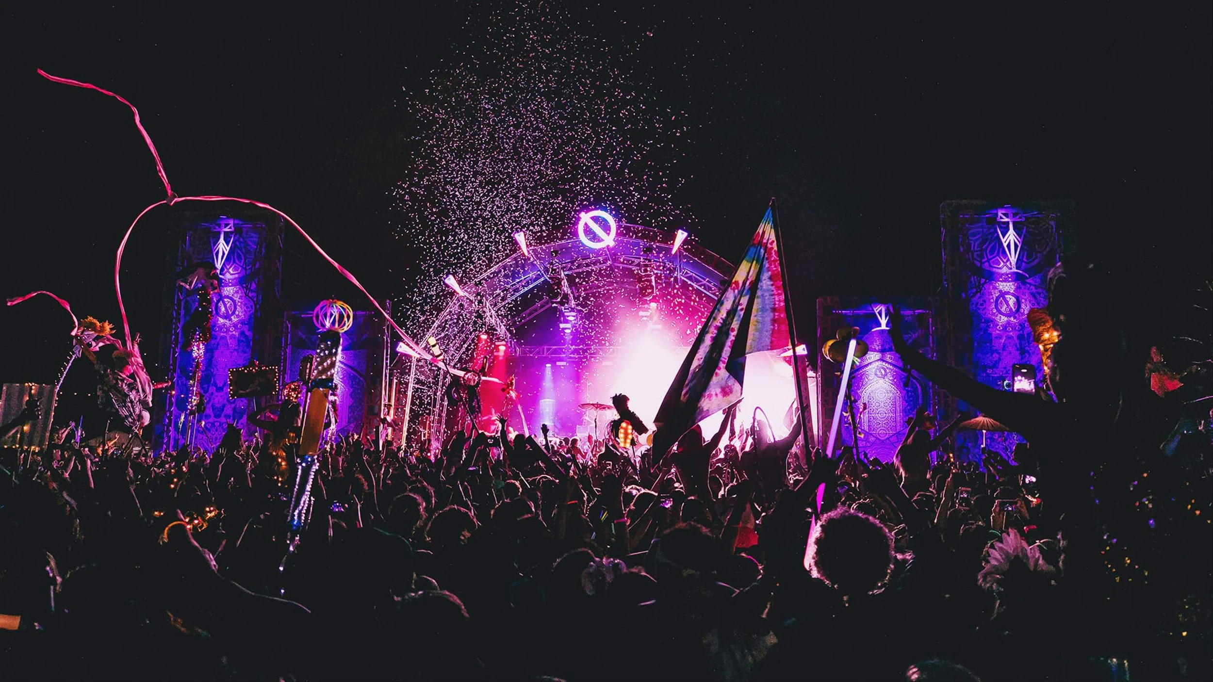 Crowd at a vibrant outdoor concert at night with fireworks and colorful stage lighting.