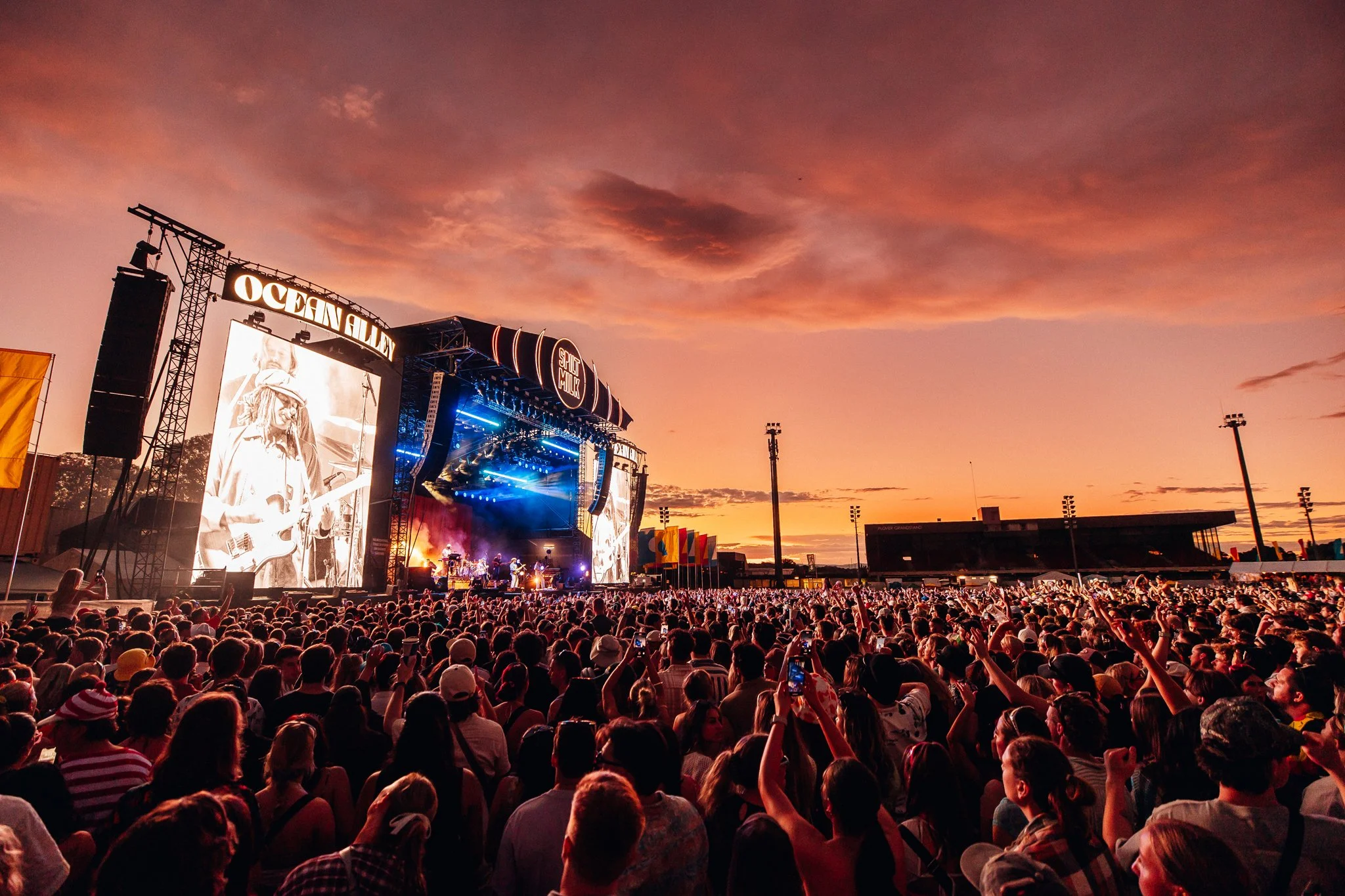 Large crowd attending an outdoor concert at sunset, with a stage, large screens, and various instruments and performers performing.