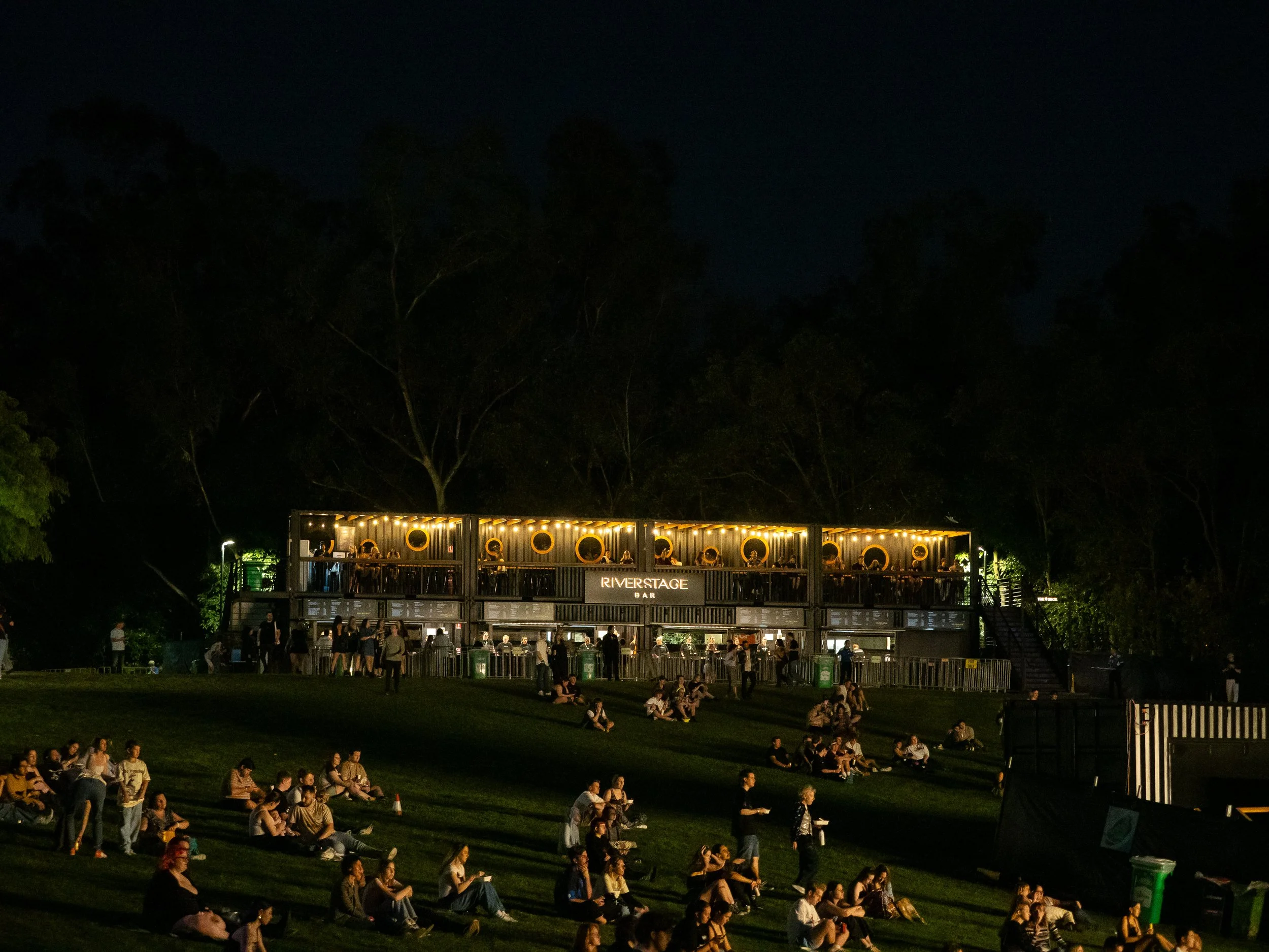 People sitting on a grassy hill at night, watching a movie projected on a large outdoor screen, with a two-story bar structure labeled 'Riverstage Bar' illuminated in the background.