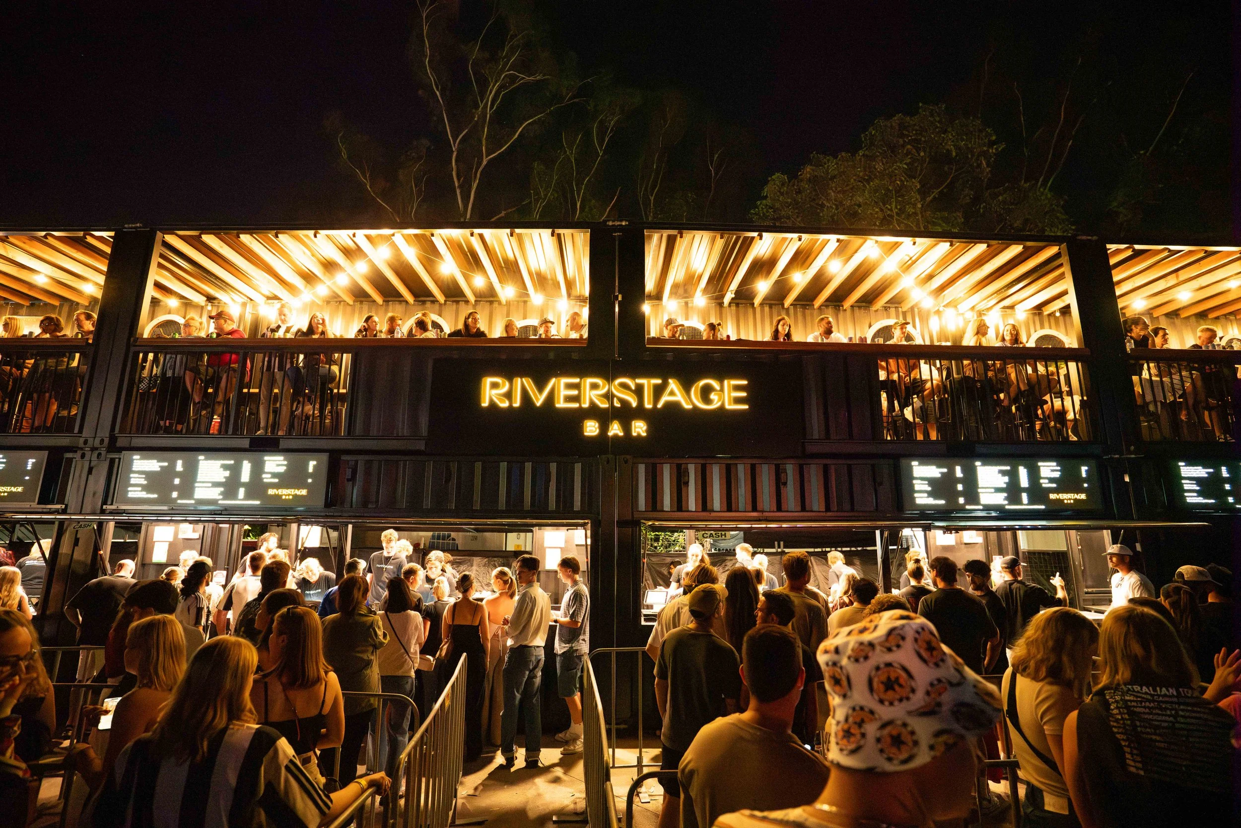 Night view of the Riverstage Bar with people standing in line and sitting upstairs, illuminated by warm yellow lights, with trees visible in the background.