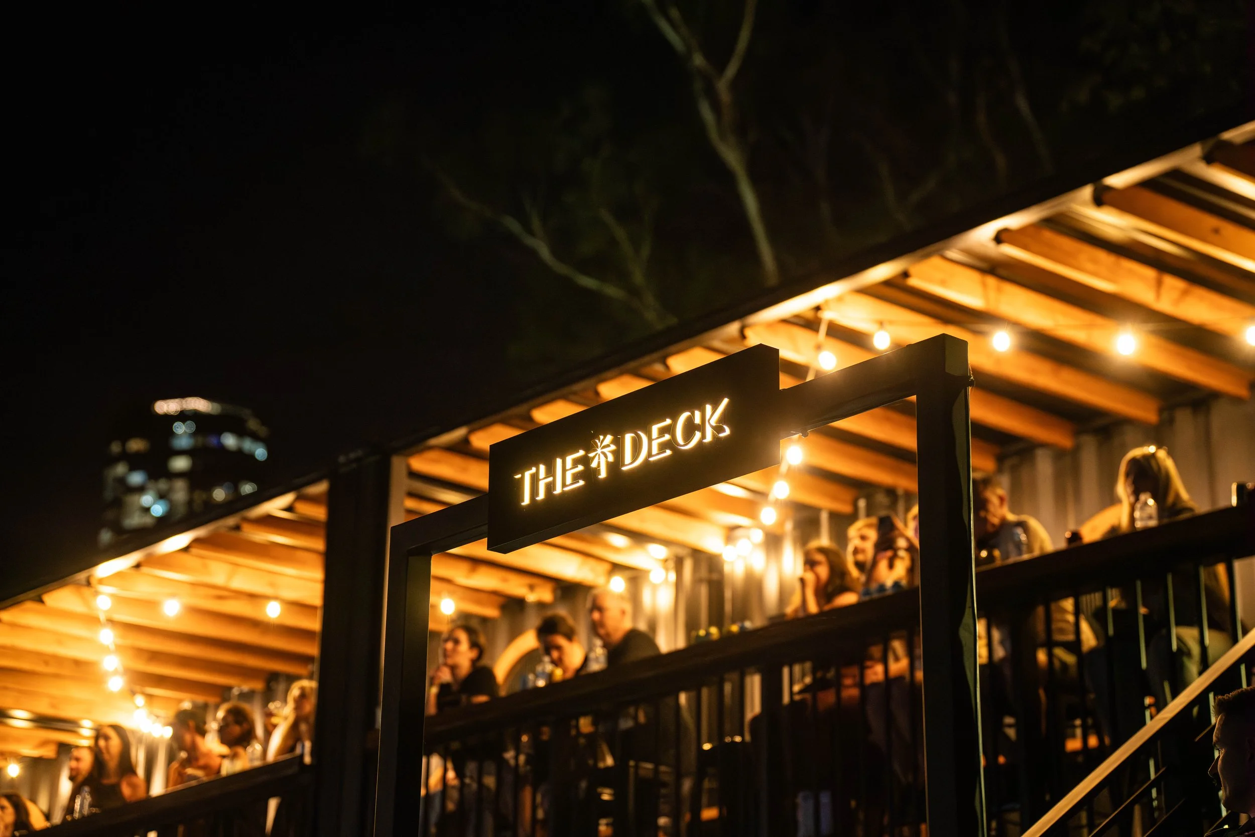 Night scene at outdoor bar named 'The Deck' with people sitting under a wooden overhang decorated with string lights.