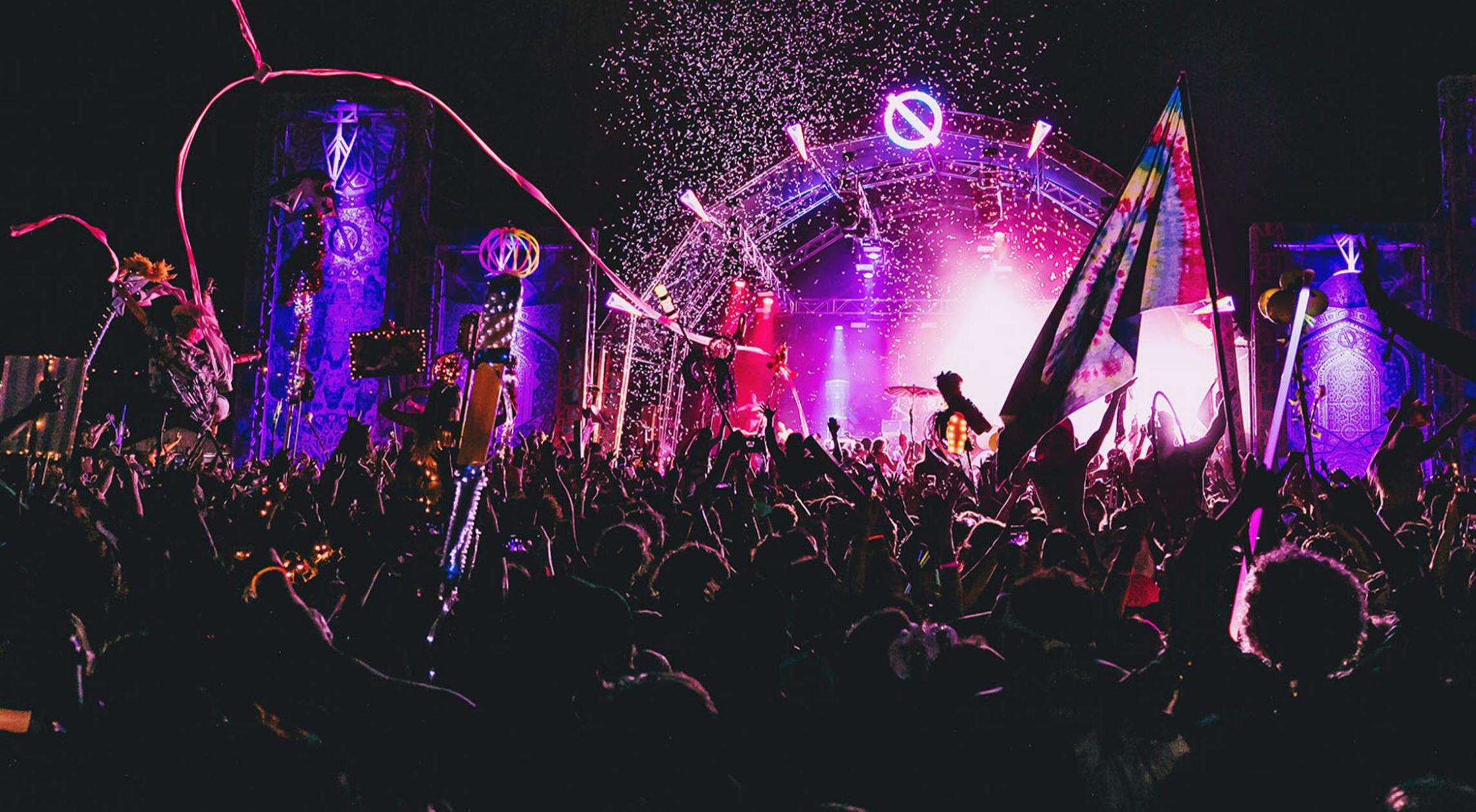 Crowd dancing at a vibrant concert with colorful lights, flags, and confetti.