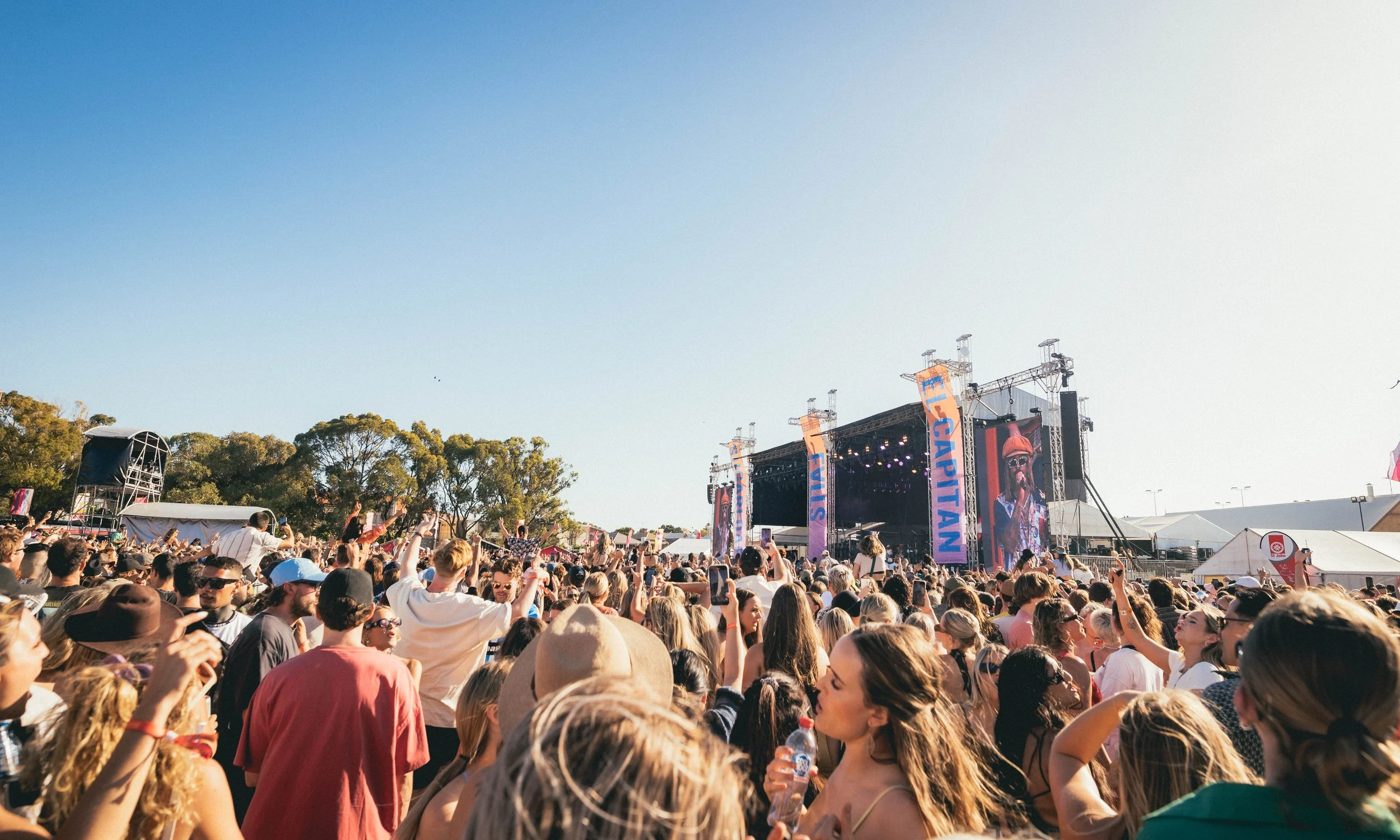 Crowd of people in front of stage during the day with blue skies and trees in the background. Spilt Milk Perth.