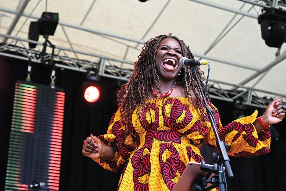 Woman of colour with curly hair in vibrant outfit singing at the Australia Day concert.