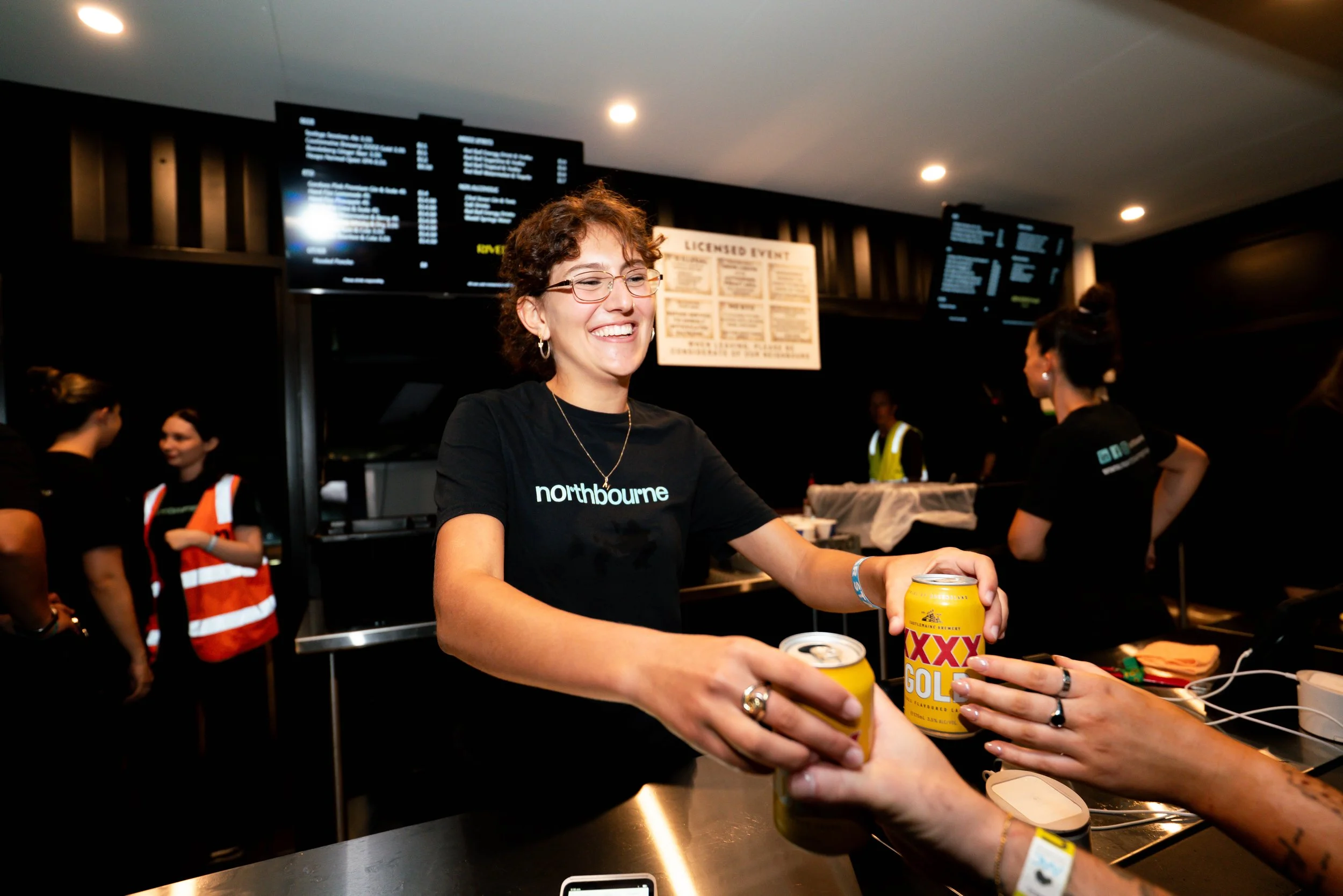 A woman wearing glasses and a black shirt with 'northbourne' on it is smiling and handing a can of XXXX Gold beer to someone at a counter in a busy indoor setting, with other staff and screens in the background.
