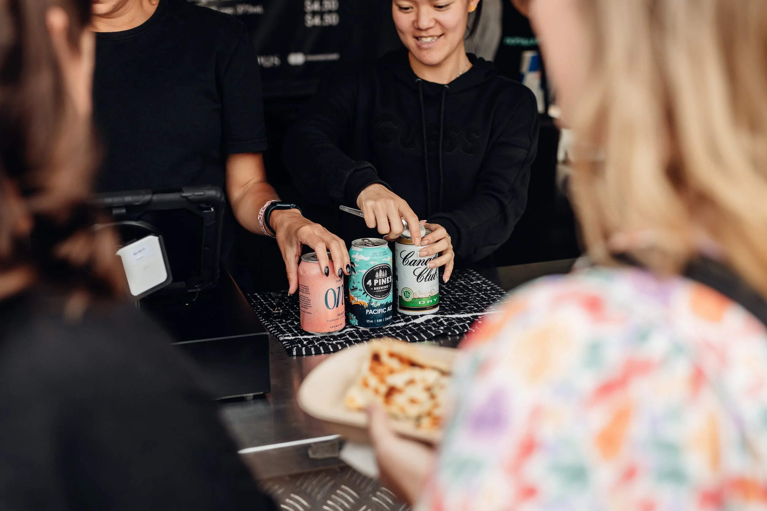 A group of people at a counter ordering or paying for drinks, with a person in a black hoodie serving canned beverages, and someone in the foreground holding a plate of pizza.