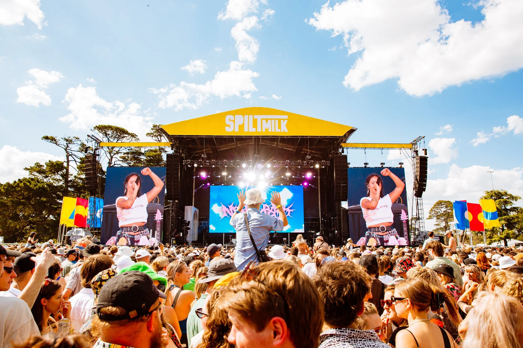 Crowd of people attending an outdoor concert at the Split Milk music festival with a large stage, big screens, and flags, under a partly cloudy sky.