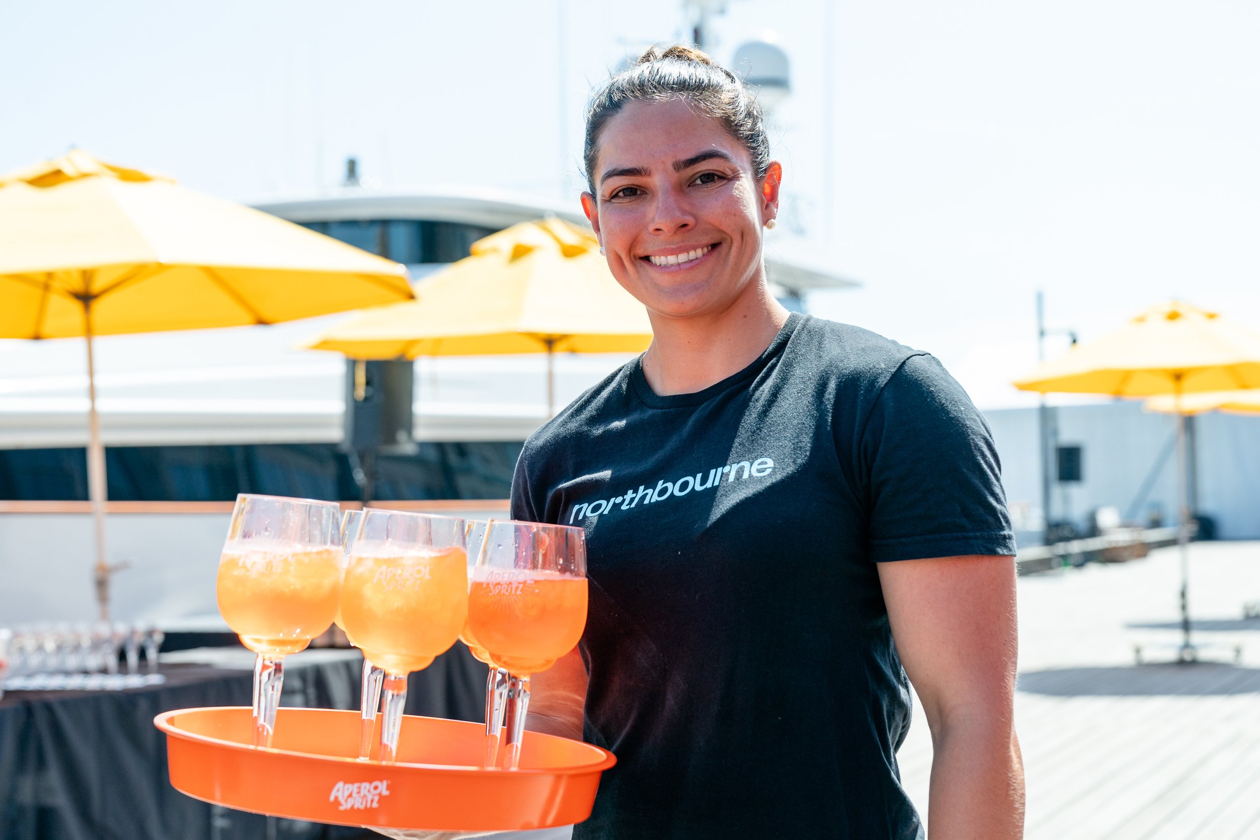 Girl in black northbourne branded top carrying tray of aperol spritz glasses.