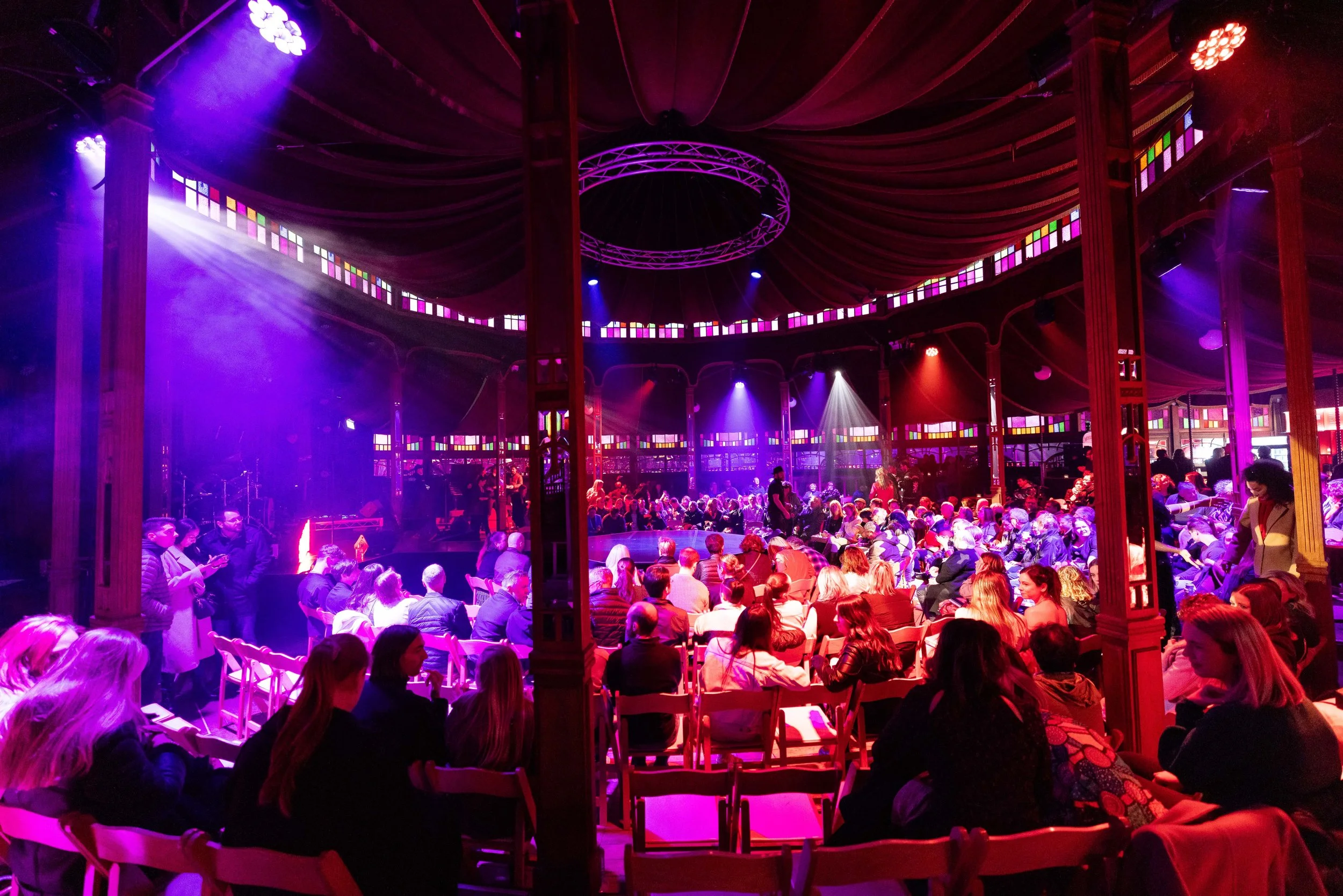 An indoor theater with colorful stage lights illuminating an audience seated in chairs, waiting for a performance to begin.