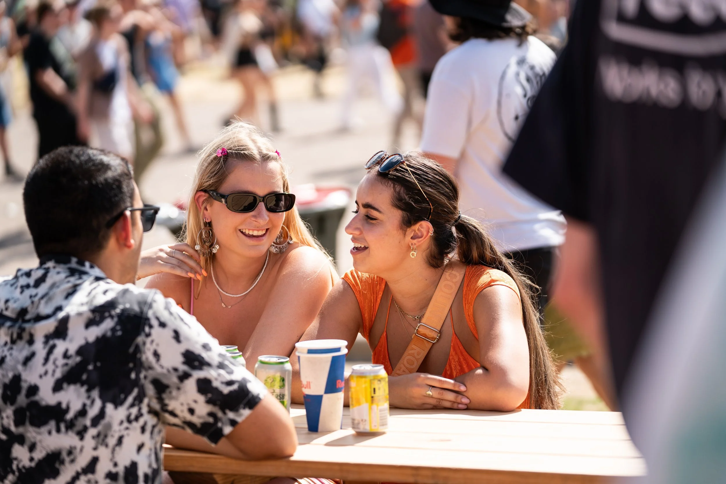 3 young adults sitting at a festival table, sharing canned drinks.
