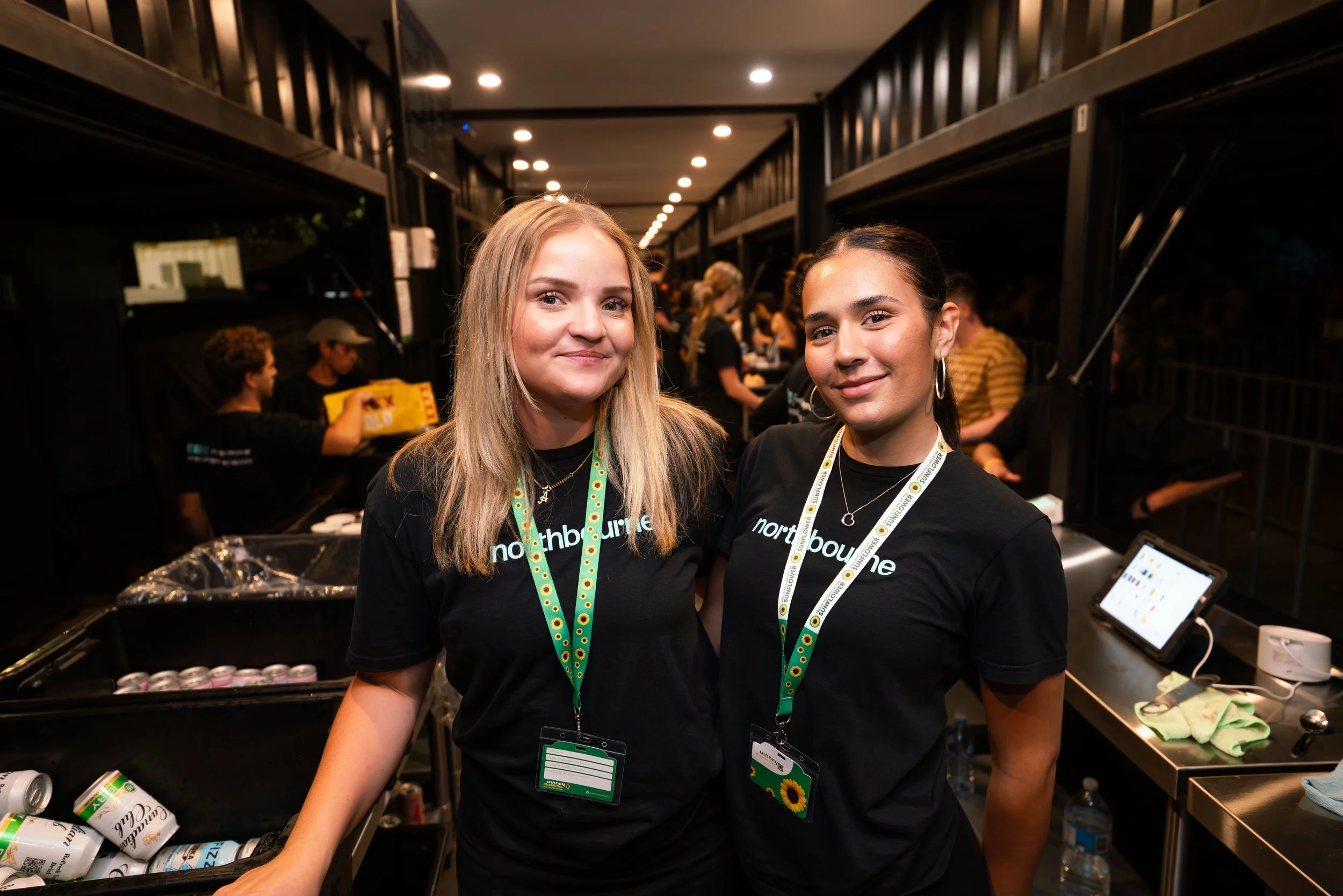 Two young women with name tags and black t-shirts standing together at an event, smiling at the camera, with a busy background of people and equipment.