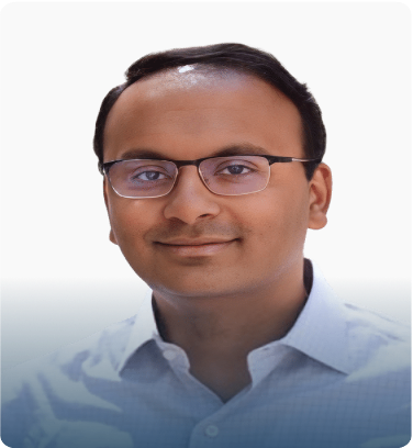 Portrait of a man with glasses wearing a light blue shirt, smiling against a gray gradient background.
