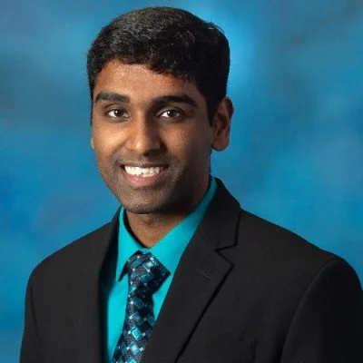 Professional headshot of a young man in a black suit, turquoise shirt, and patterned tie, smiling against a blue background.
