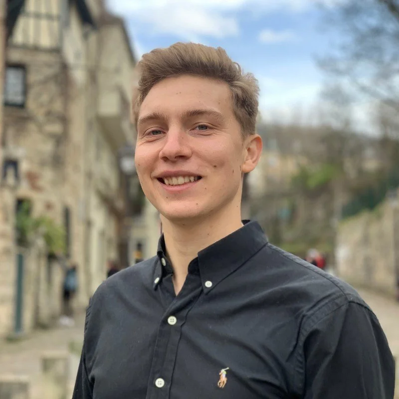 Young man smiling outdoors in front of historic buildings during daytime, wearing a black button-down shirt.