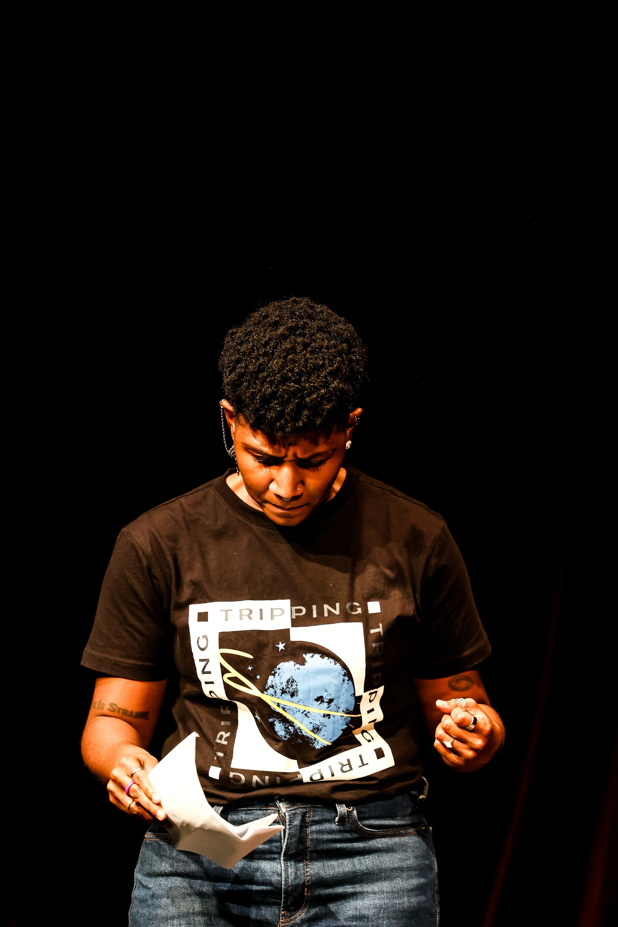Kenyatta looking down on a stage. They are standing in front of a black background. Their hands are out in fronf of them, gripping a page. Kenyatta's eyebrows are furrowed.