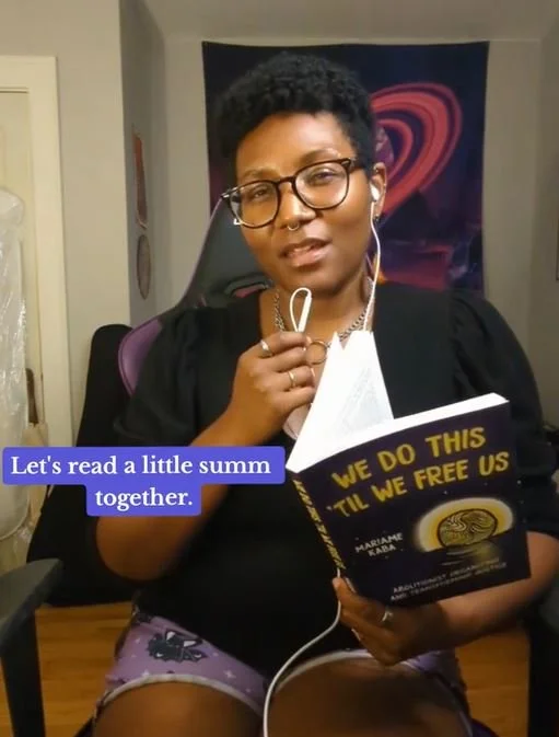 Kenyatta looking into the camera. She's holding a book that reads "We do this till we free us". There is text on the screen that reads "Let's read a lil summ together."