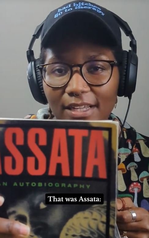 Kenyatta looking into the camera. She is holding a book that reads "Assata"
