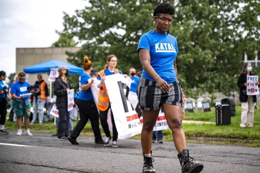 Kenyatta looking straight ahead while walking in the street. They are wearing a blue shirt, shorts, and combat boots. Image by Tyler Russel, CT Public, 2022